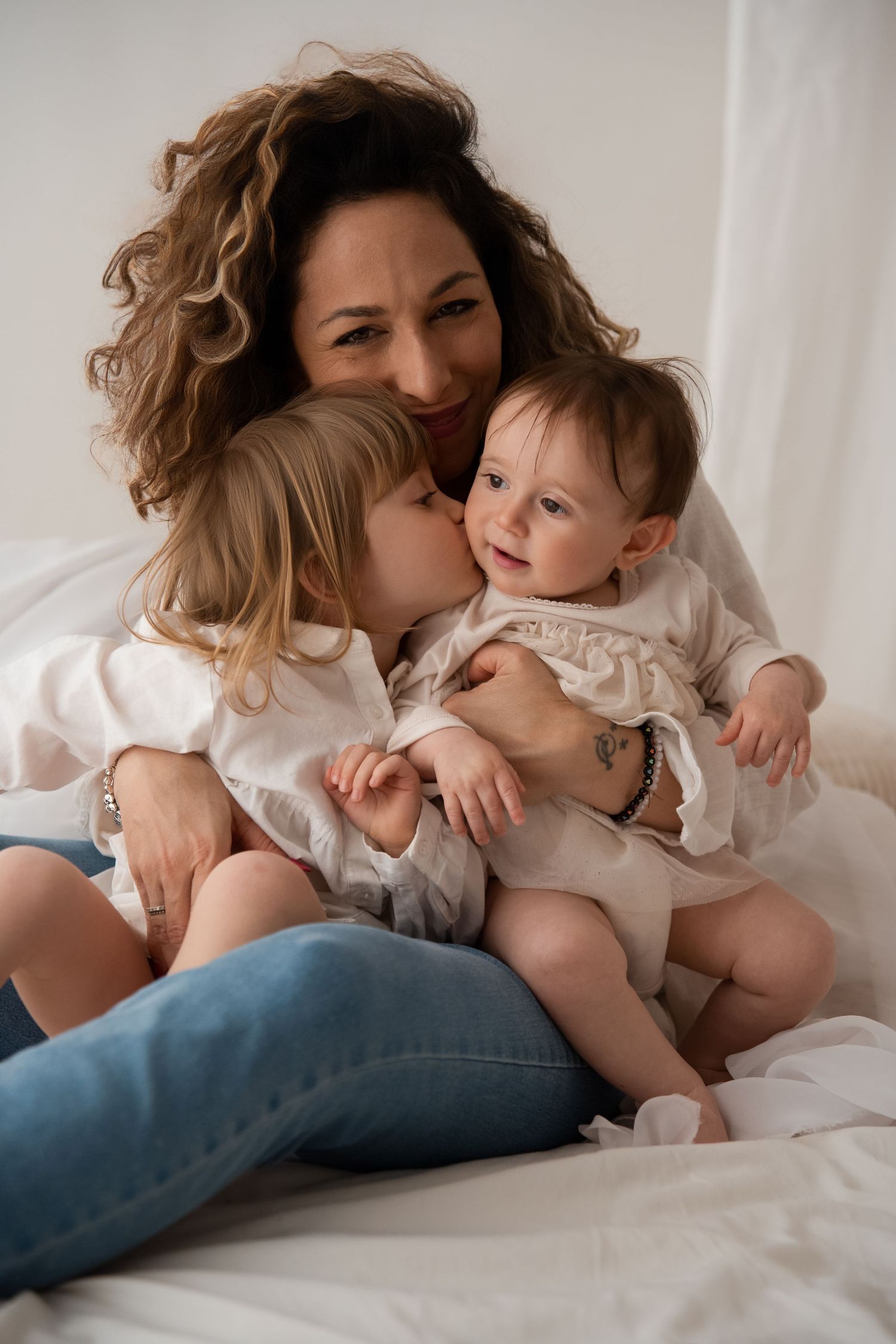 A curly-haired mother smiles while holding her baby as her older daughter gives a kiss.