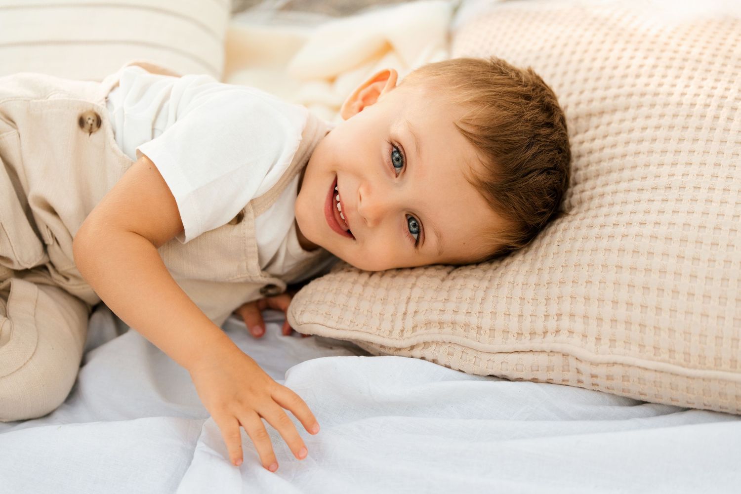 An adorable smiling toddler boy relaxes on a bed with beige pillows.