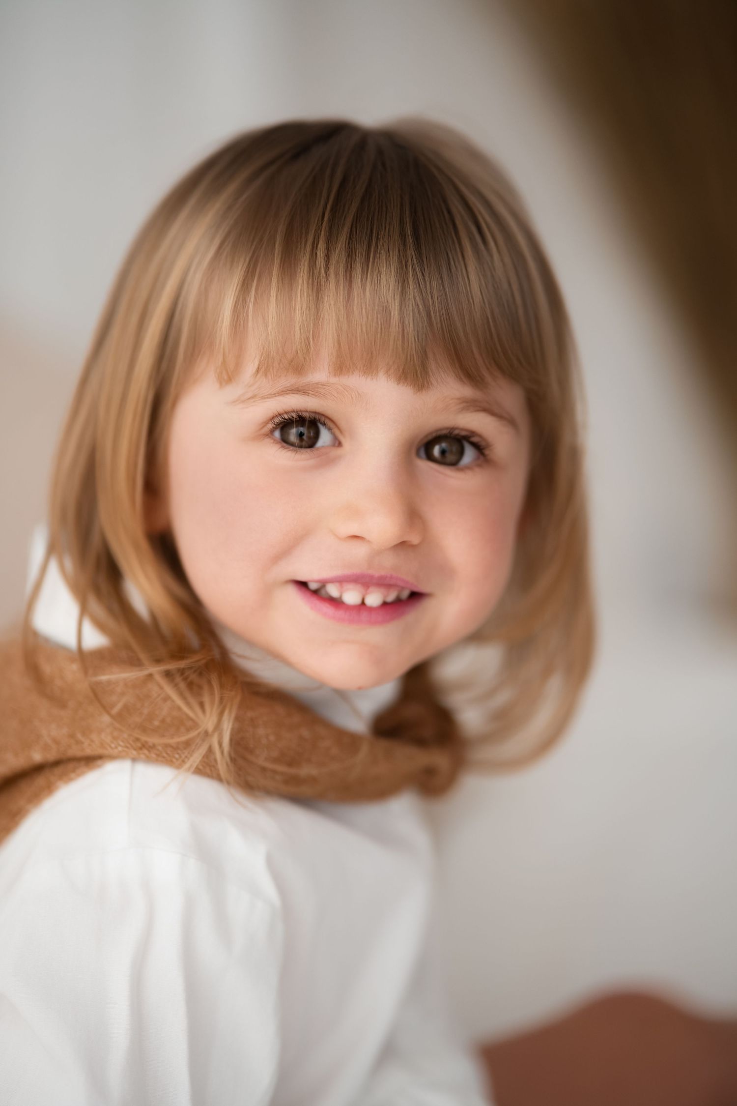 A cute young girl with blonde bangs and brown eyes smiles warmly at the camera.