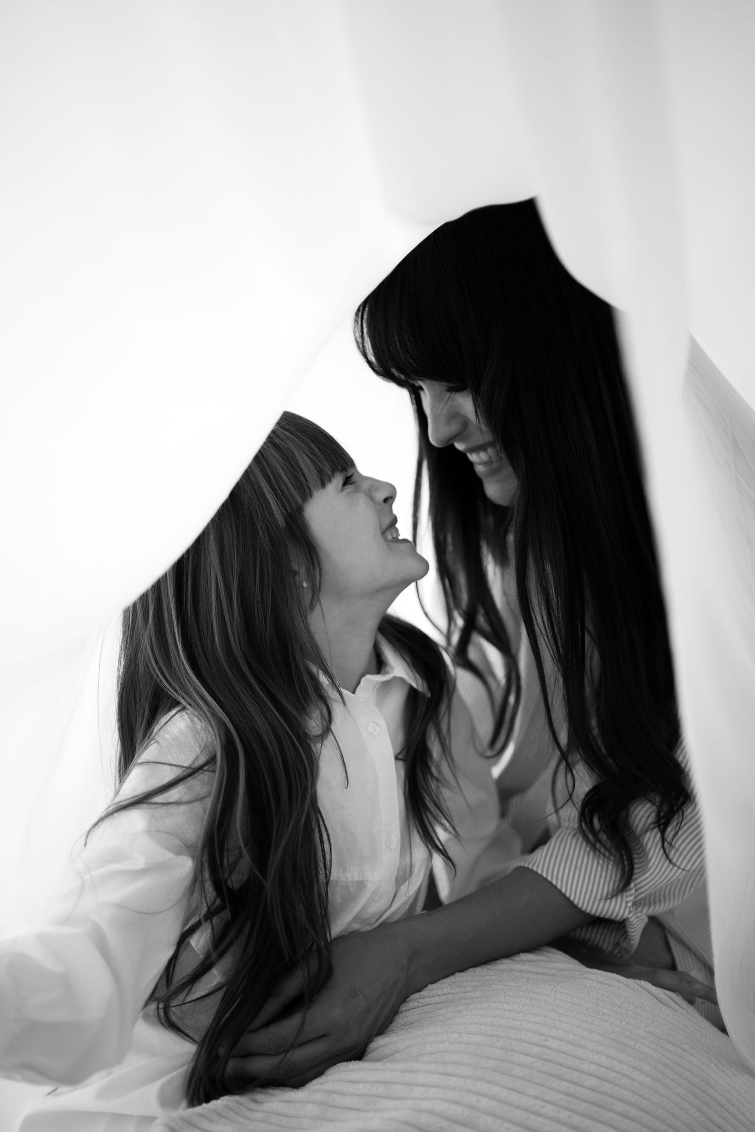A mother and daughter with long dark hair smile at each other under a white sheet.