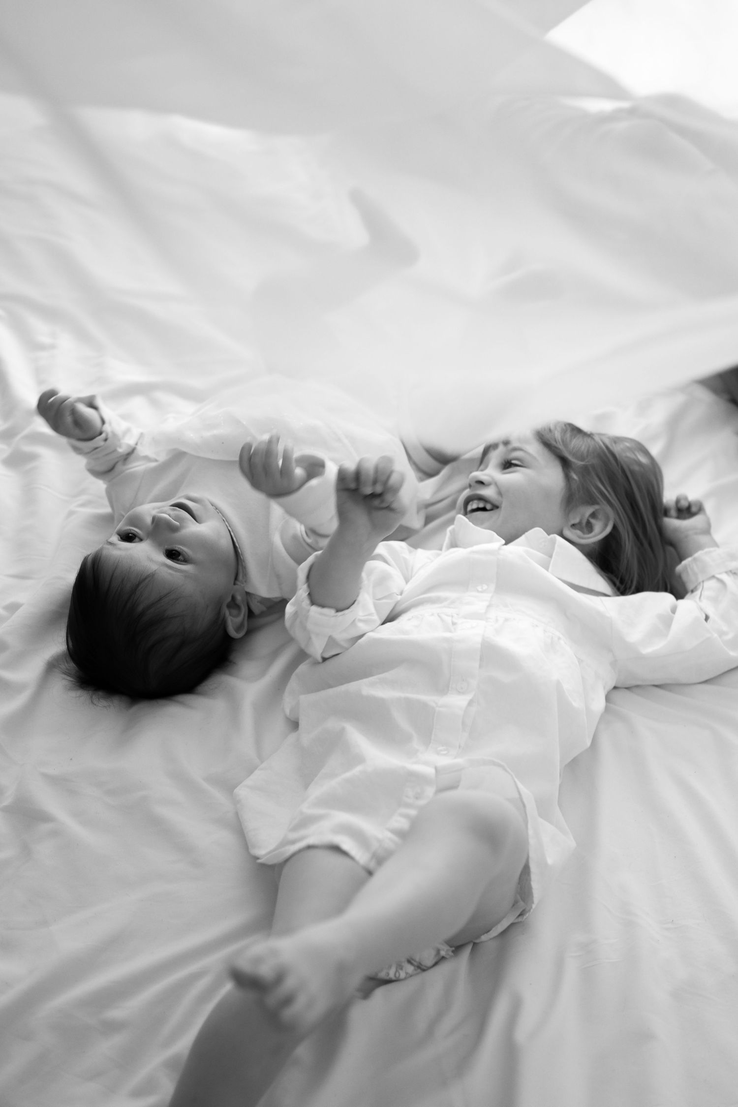 A black and white photo of a baby and young girl laughing together on white bedding.