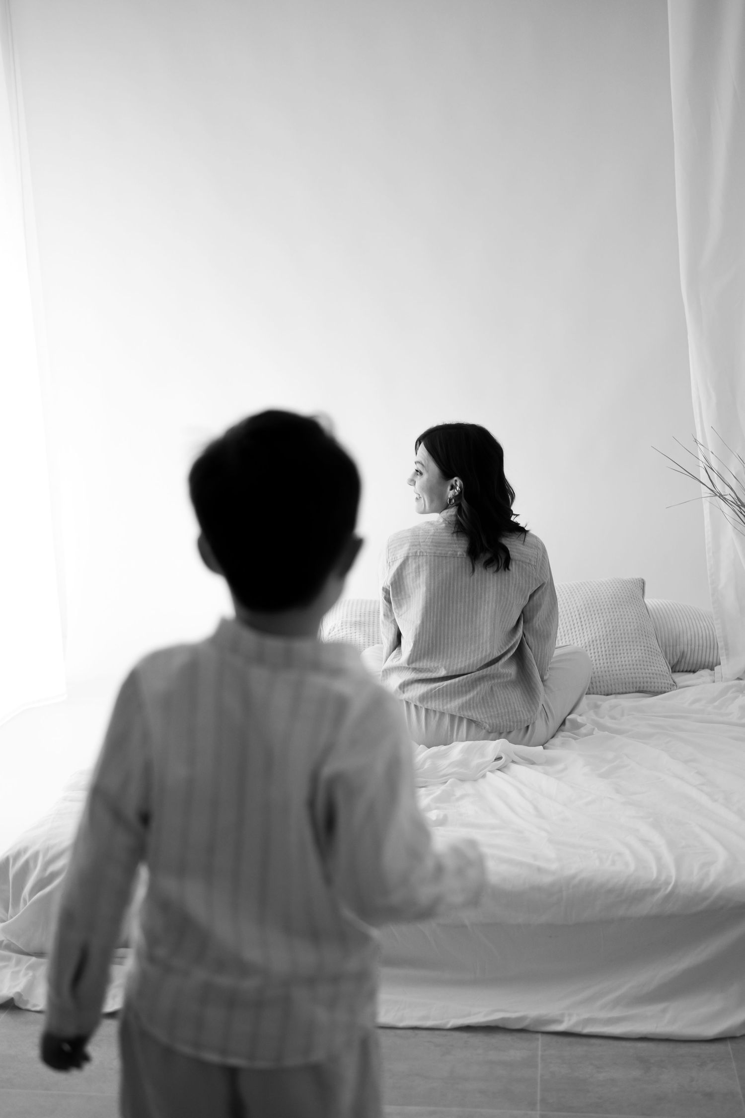 Black and white photo of child approaching mother sitting on bed in bright white room.