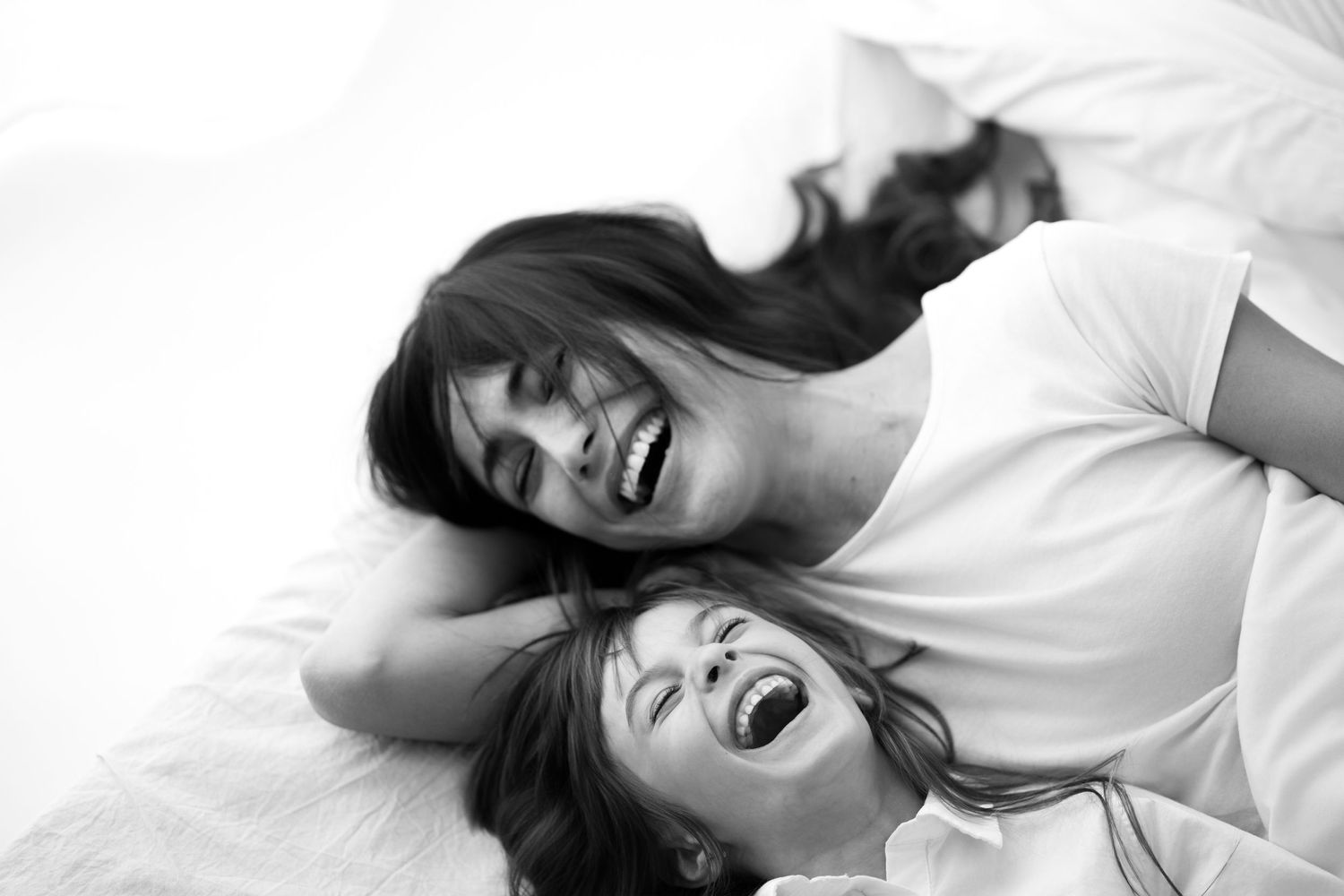 Black and white photo of mother and daughter laughing joyfully on white bed.