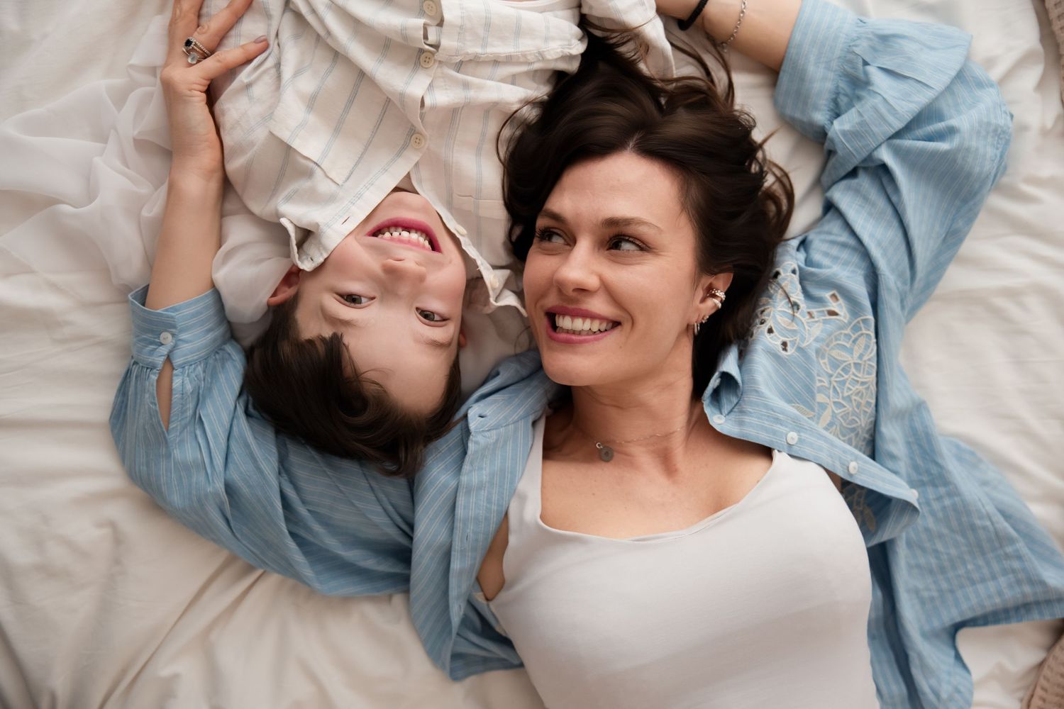 Happy mother and young daughter lying together on white bed, smiling at each other.