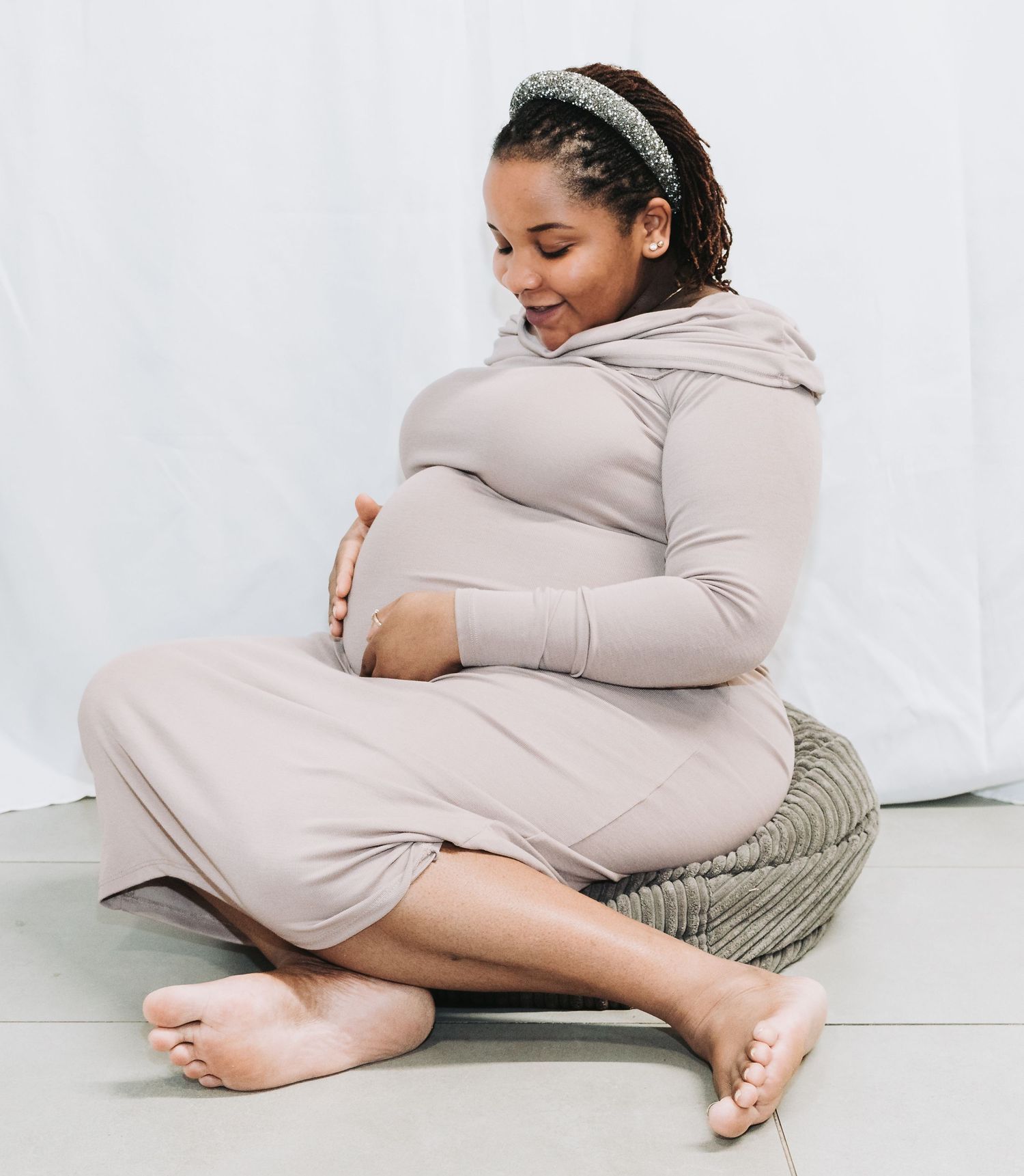Expectant mother in a beige dress sits comfortably on floor cushions while cradling her baby bump.