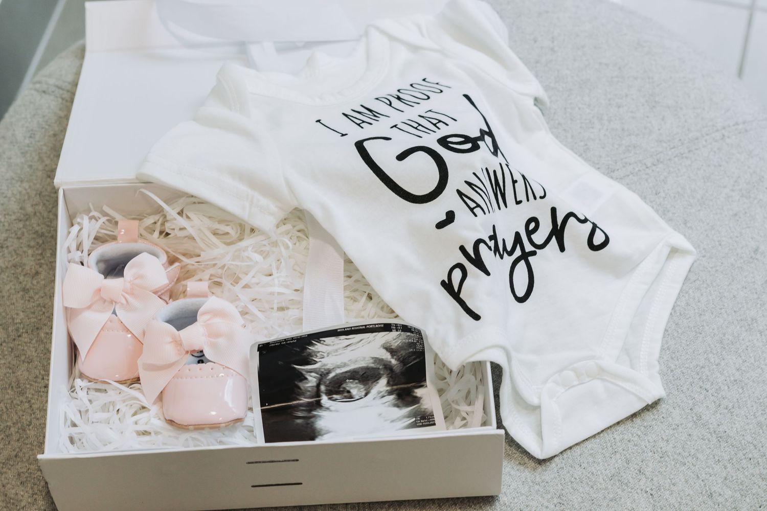 Box containing baby onesie with text, pink baby items, and a black and white sonogram photo.