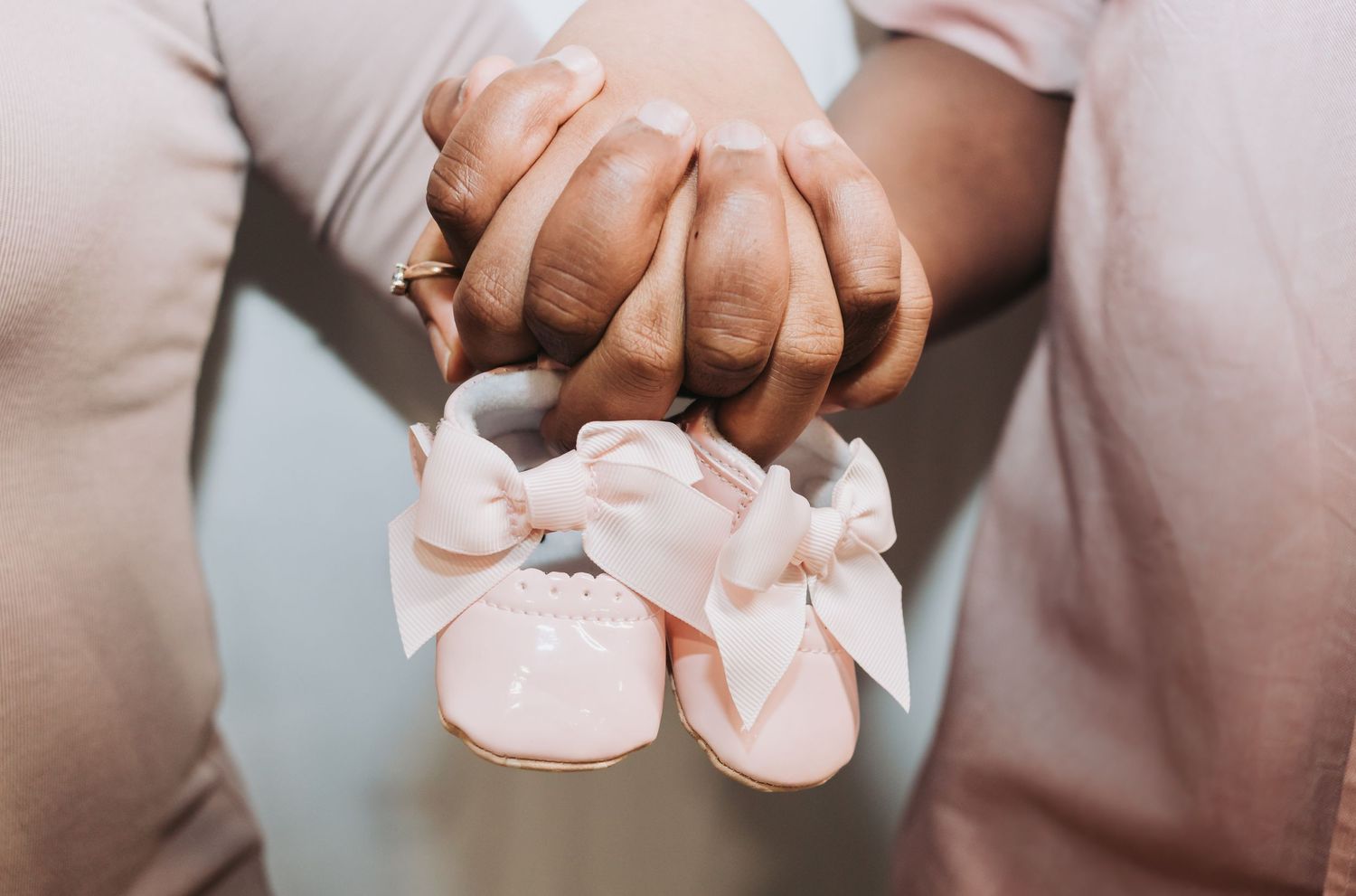 Hands holding tiny pink baby booties with bows against a soft neutral background.
