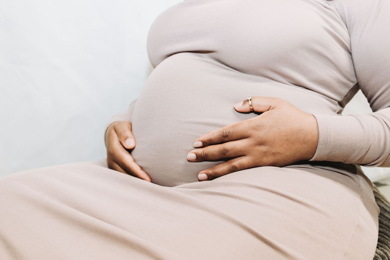 A pregnant person wearing beige clothing holds their belly with both hands against a light background.