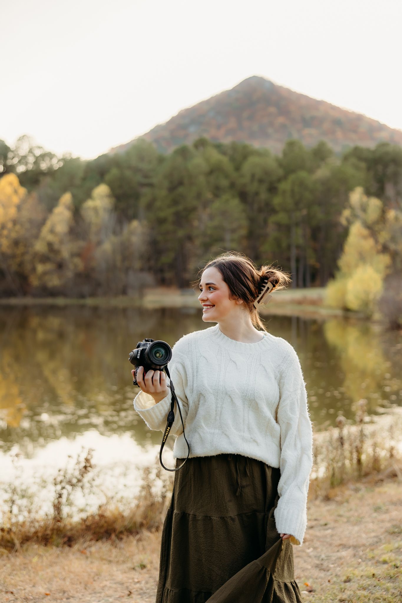A photographer in a white sweater and dark skirt stands by a lake during autumn capturing scenic shots in nature.