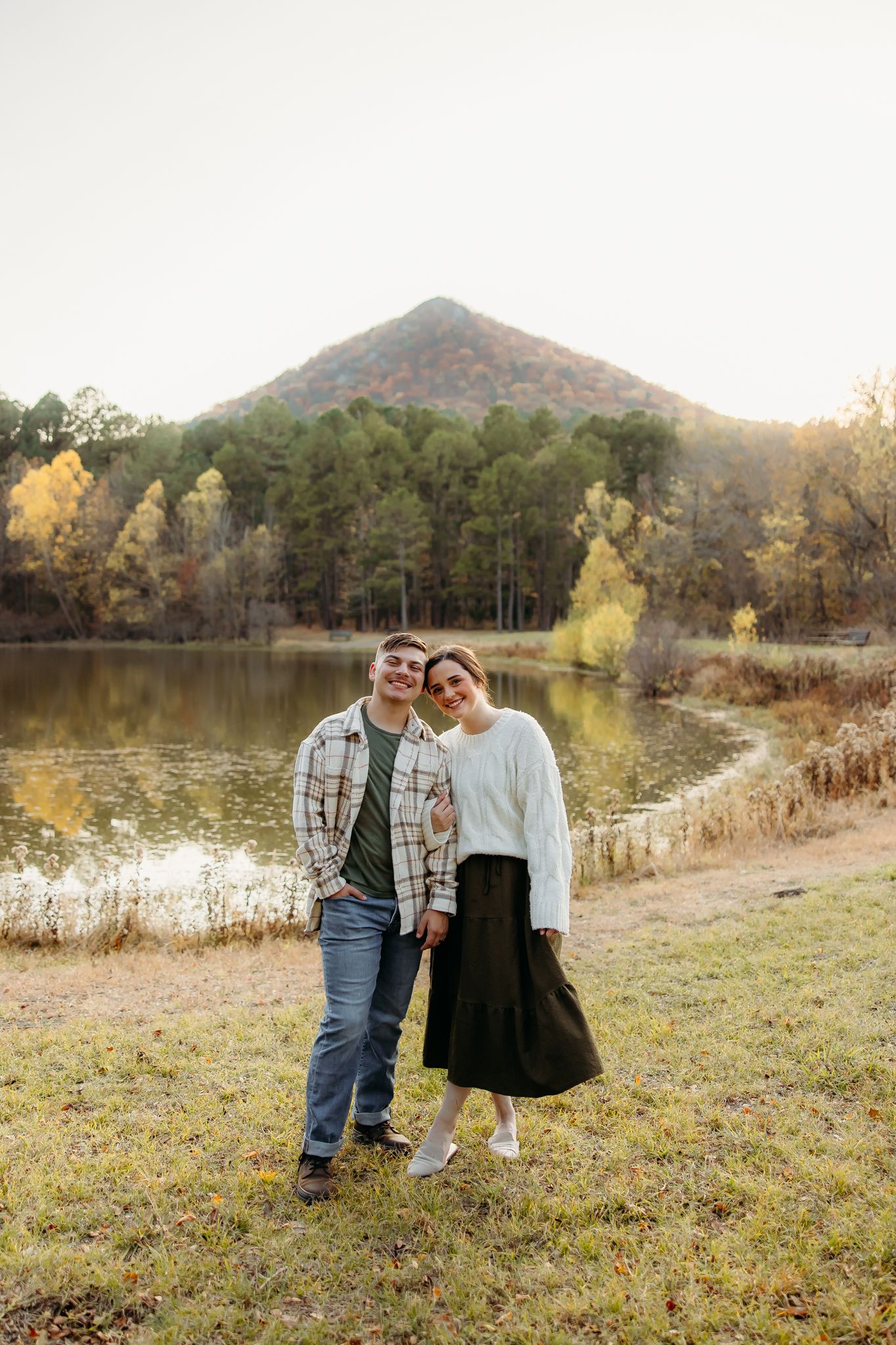 A couple stands together on grassy hill beside an autumn lake with trees and mountains in the background.