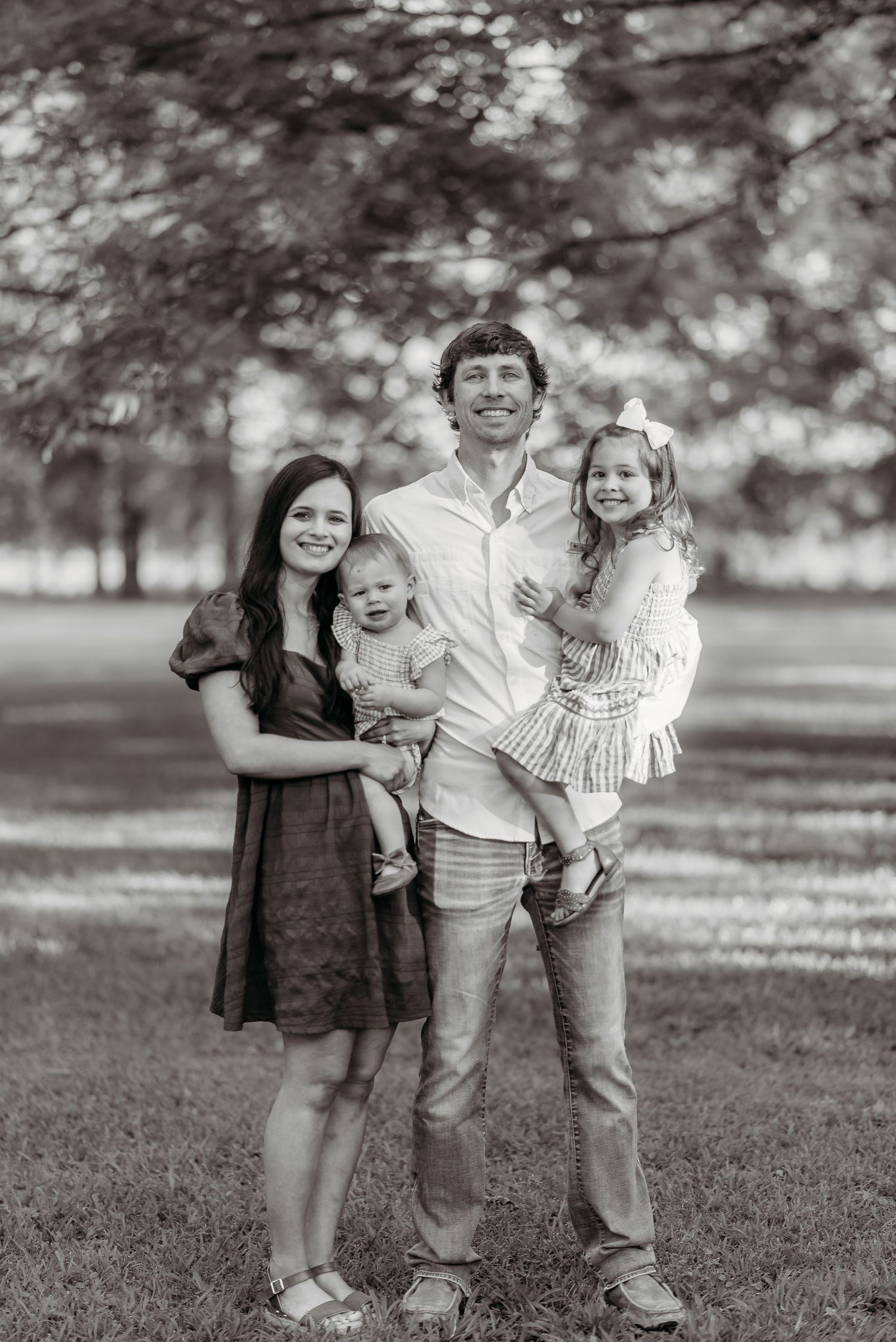 Black and white outdoor family portrait taken in a park with trees in the background.