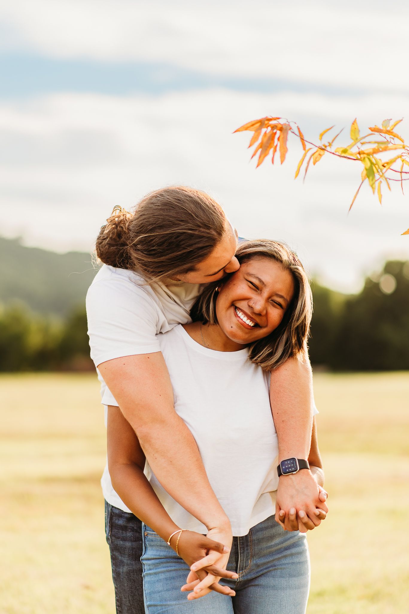 A couple in white shirts embraces while laughing in a sunny field.