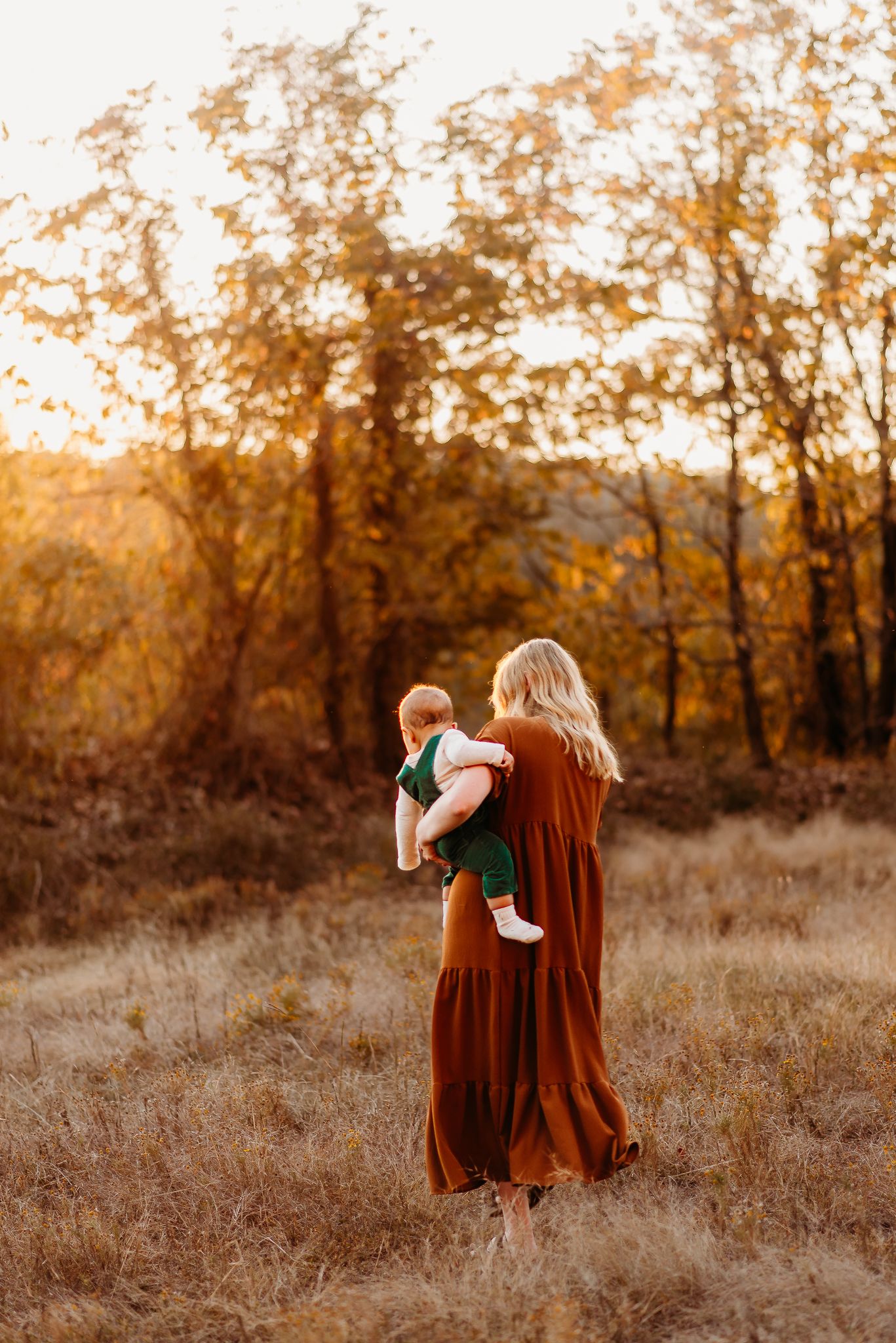 A person in a rust-colored maxi dress holds a baby against autumn trees at sunset.