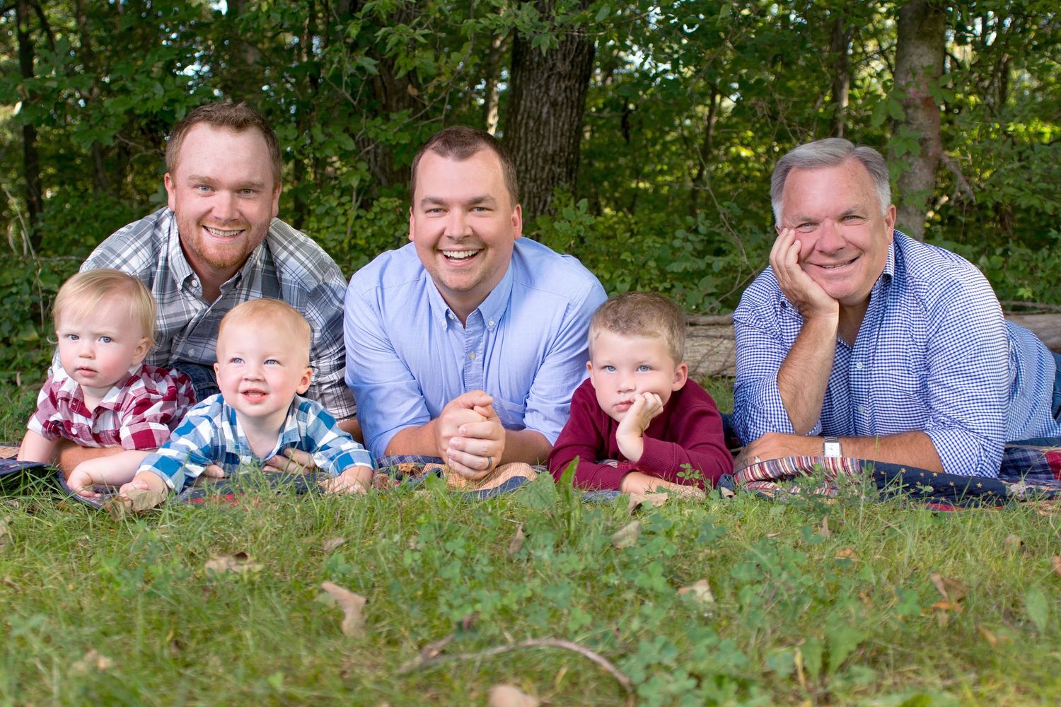 Three generations of men and two toddlers smile together outdoors on grass with trees in the background.