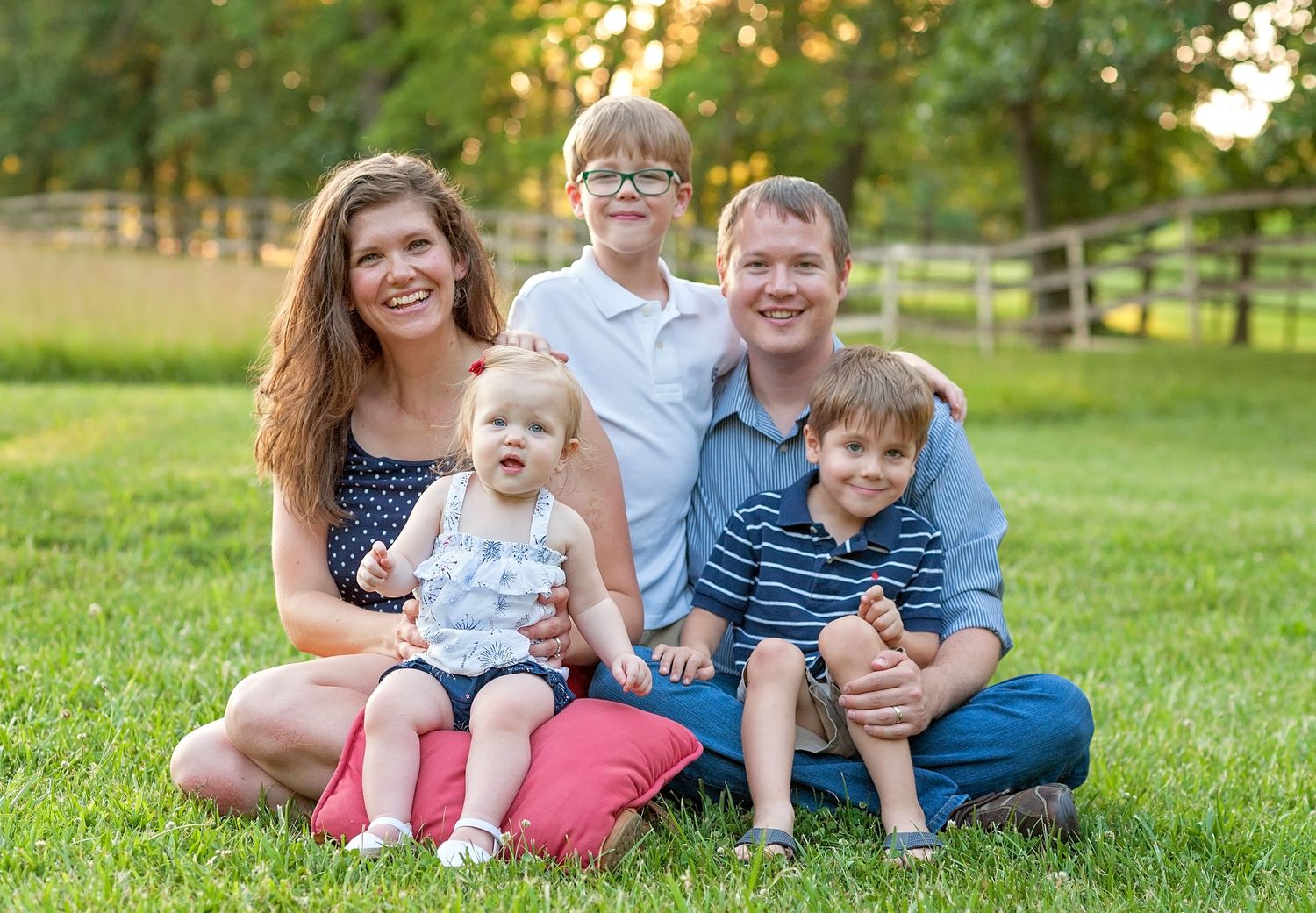 Happy family of five smiling outdoors on green grass with a wooden fence and trees in the background.