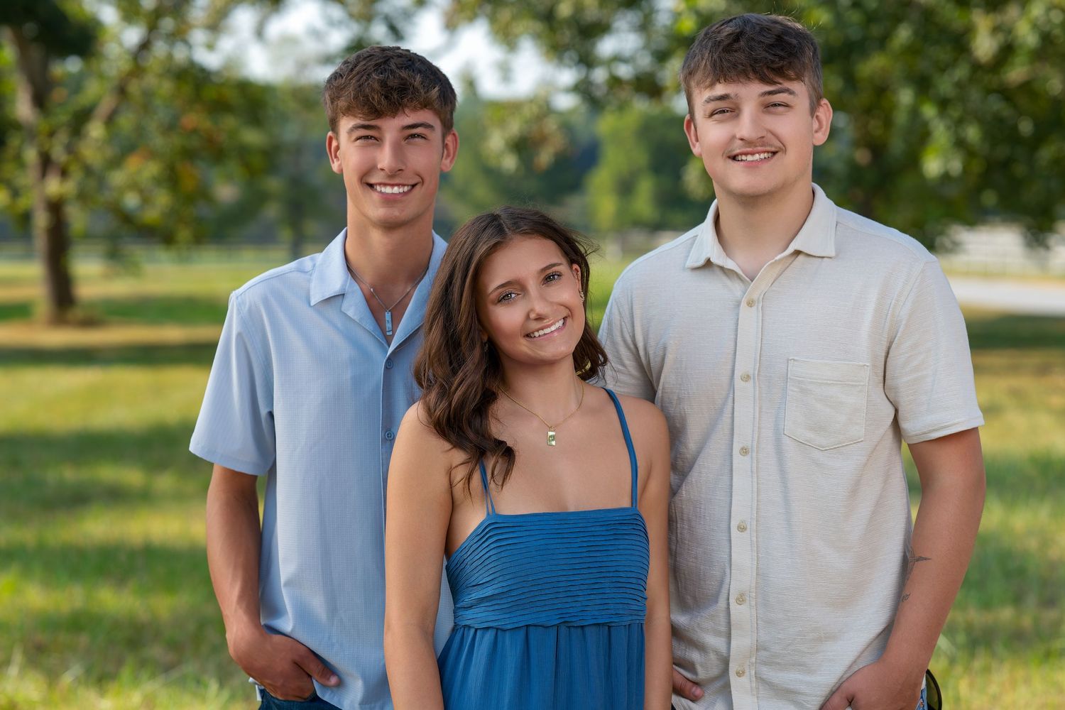 Three smiling teenagers pose outdoors in a sunny green park, two boys flanking a girl in a blue dress.