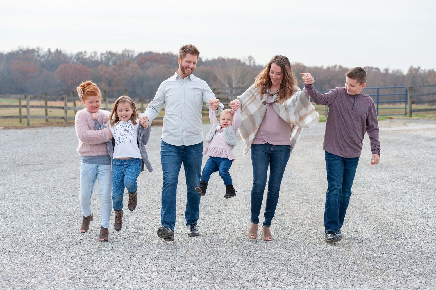 Happy family of six walking together outdoors on a gravel path, smiling and holding hands on a fall day.