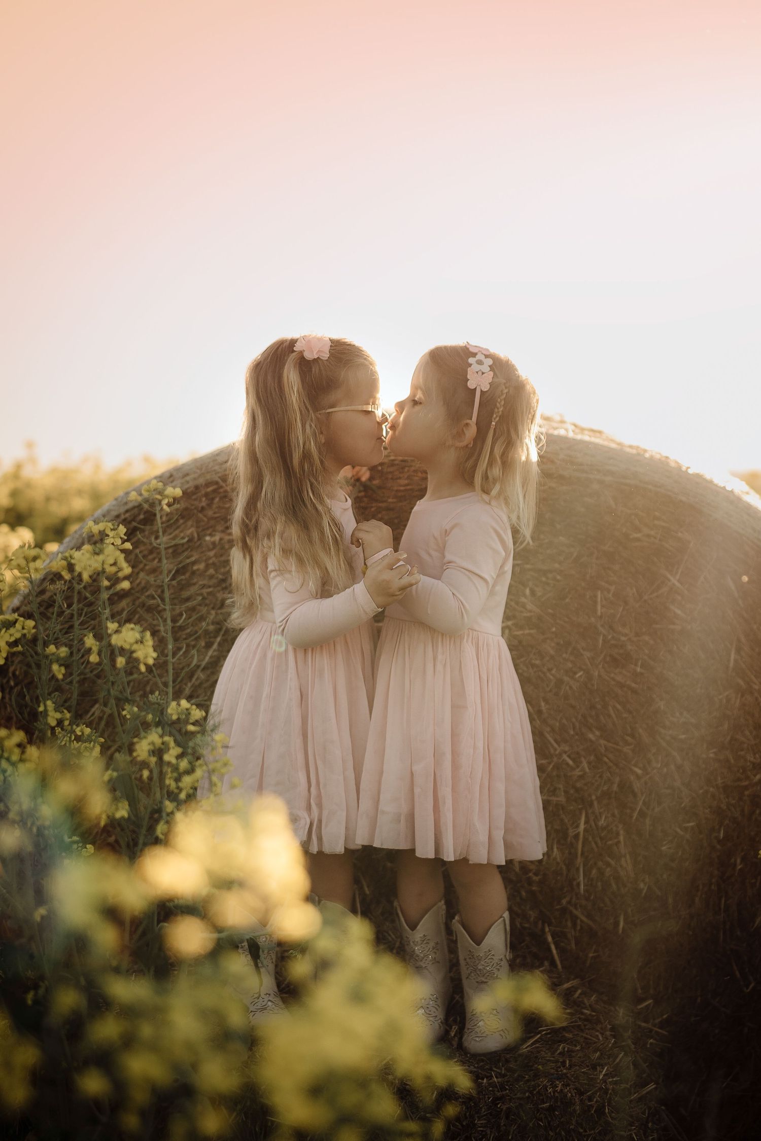 Two children in matching light pink dresses embrace in a golden sunlit field at sunset.