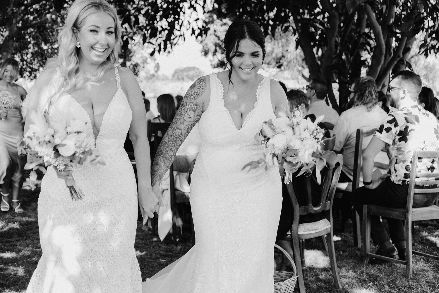 Two brides in white wedding dresses walk together holding bouquets during an outdoor garden ceremony.