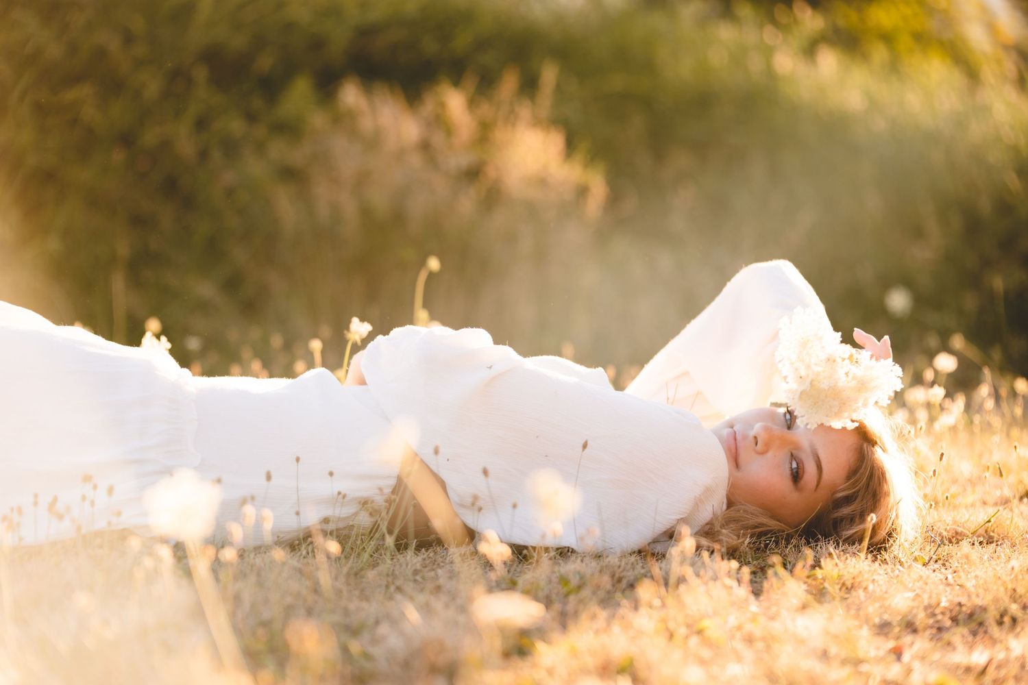 Woman in white dress lies in golden sunlit grass during outdoor portrait session.