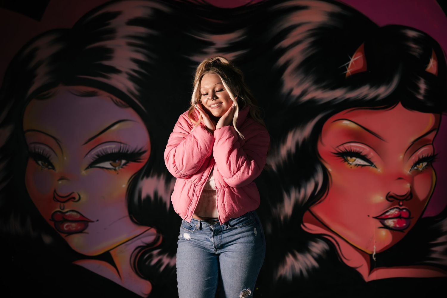 Person in a pink sweater and jeans poses in front of a vibrant wall mural featuring stylized faces with flowing hair.