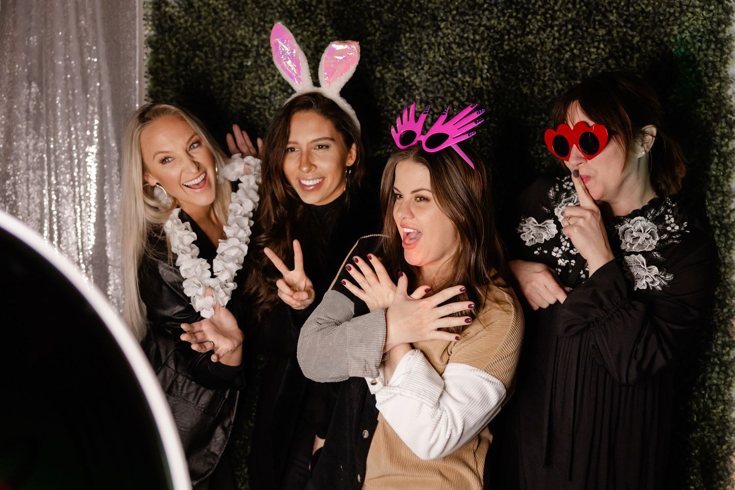 Four friends pose together in a photo booth wearing festive accessories like bunny ears and feather boas at a party.