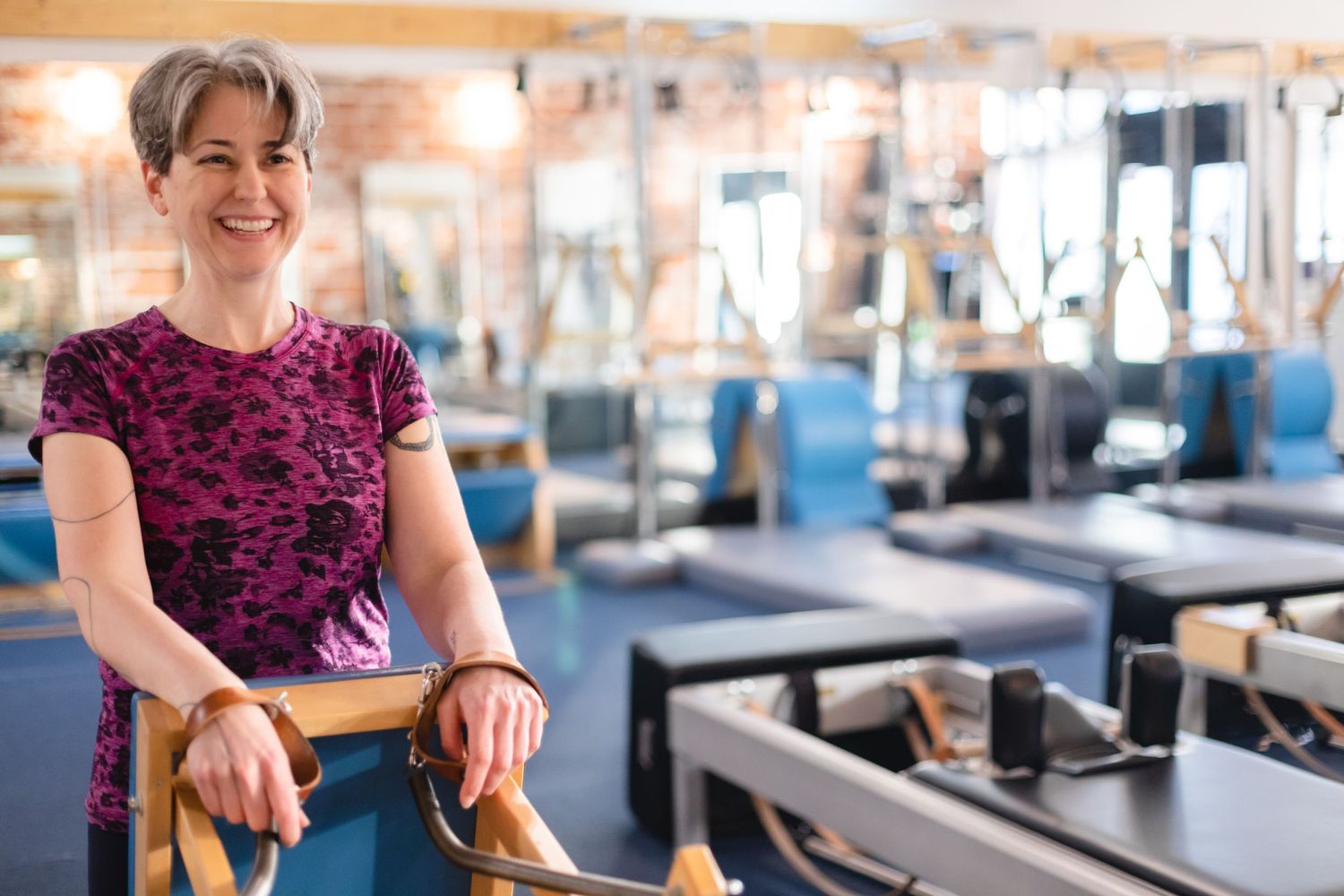 Someone in a burgundy floral top smiles while standing in a modern pilates studio with reformer equipment in the background.