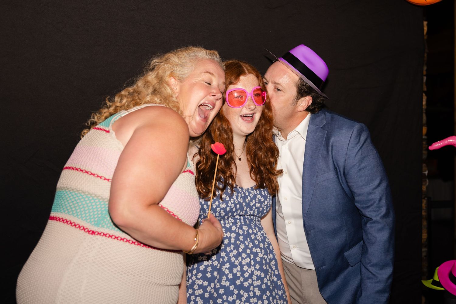 Three friends pose playfully together at a party with pink sunglasses and a purple hat.