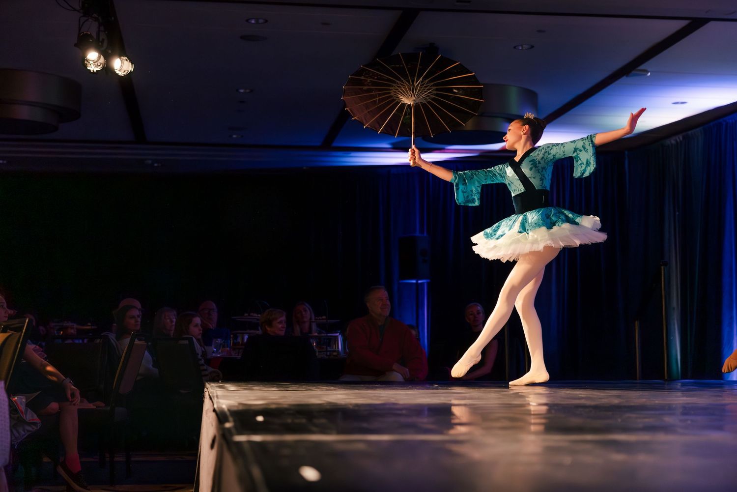 A ballet dancer in a turquoise tutu performs on stage under dramatic blue lighting.