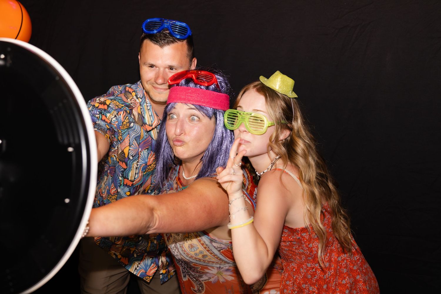 People wearing colorful outfits and novelty glasses pose playfully in a photo booth setting against a dark background.