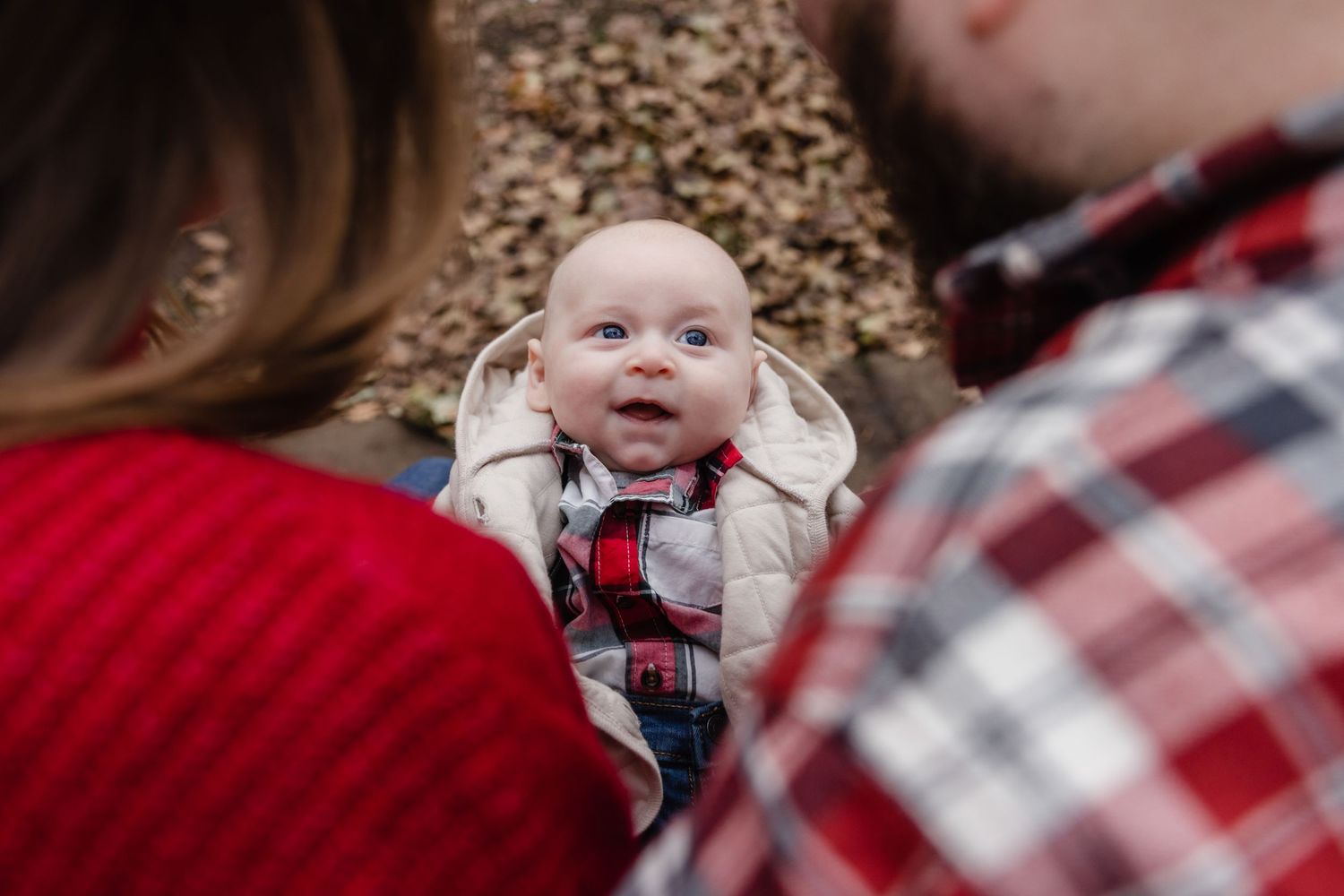 Smiling baby between two adults wearing red sweater and plaid shirt poses on autumn leaves outdoors.