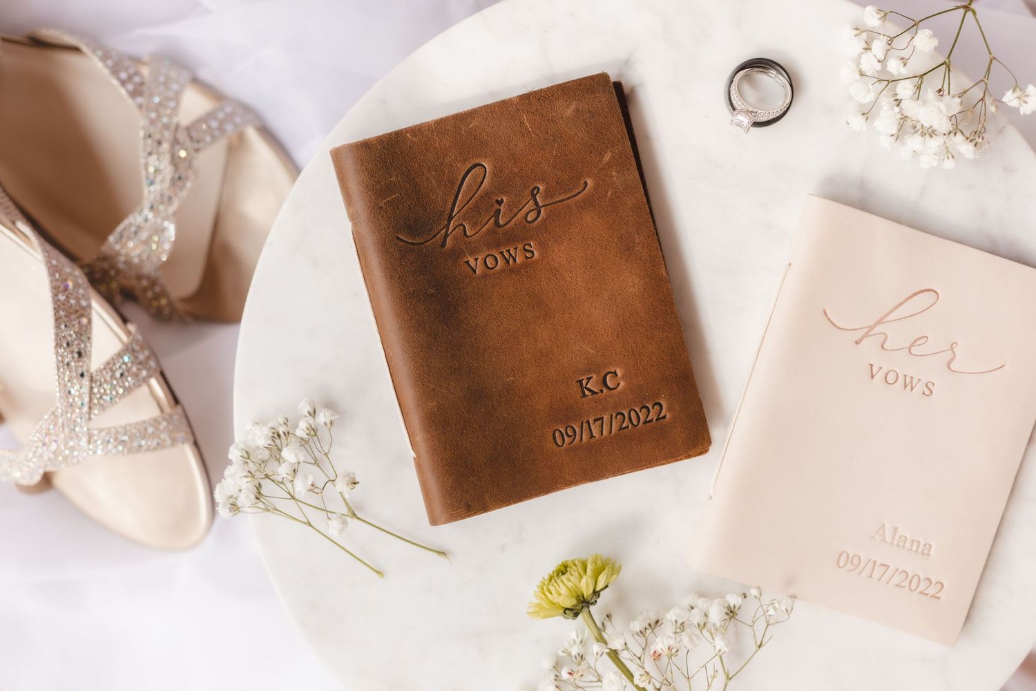 Two leather vow books with brown and blush tones displayed alongside delicate floral accents and wedding shoes.