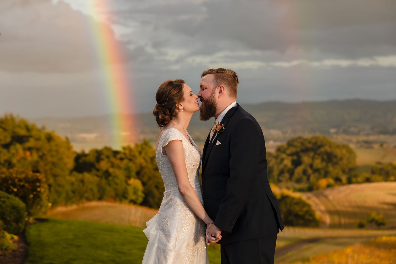 A romantic wedding moment with a double rainbow arching over rolling countryside hills during golden hour.