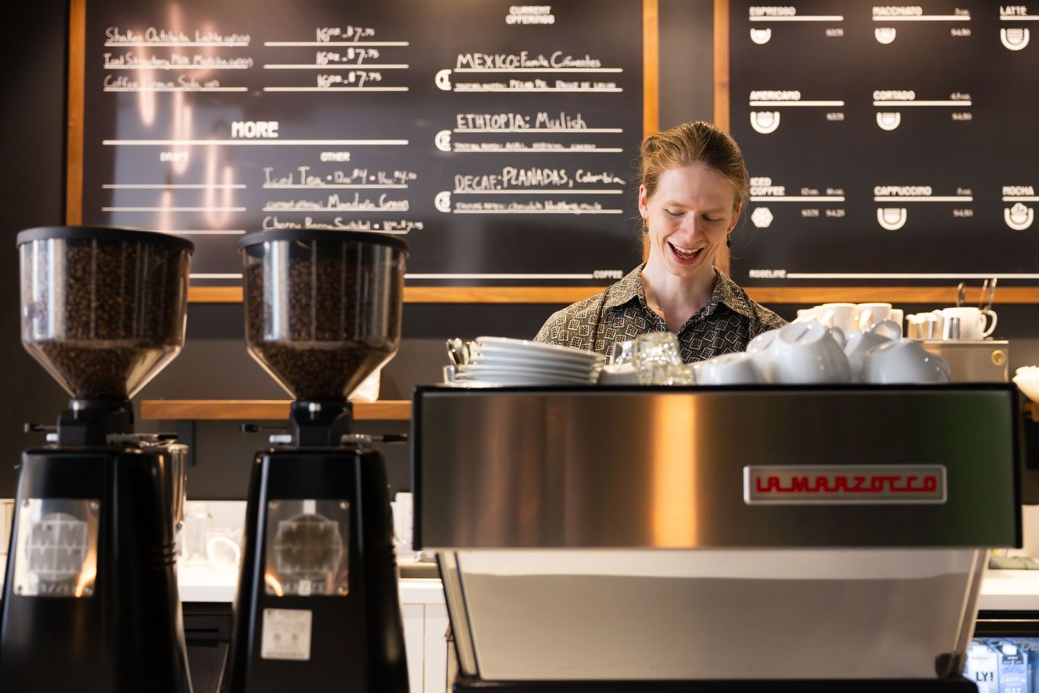 A barista stands behind an espresso machine with coffee grinders and a menu board visible in the background.