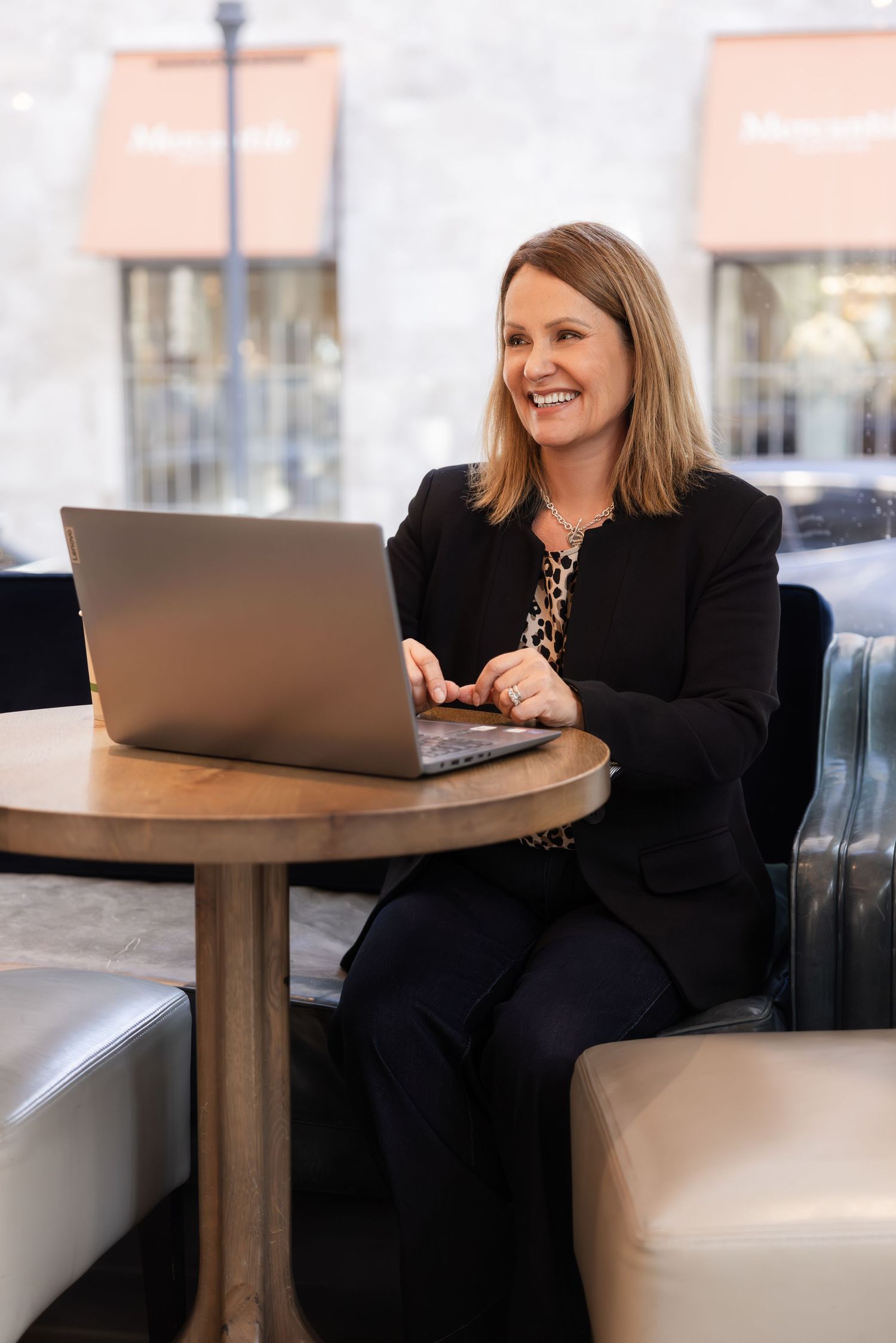 Professional woman in black blazer works on laptop while sitting at round table in modern cafe setting.