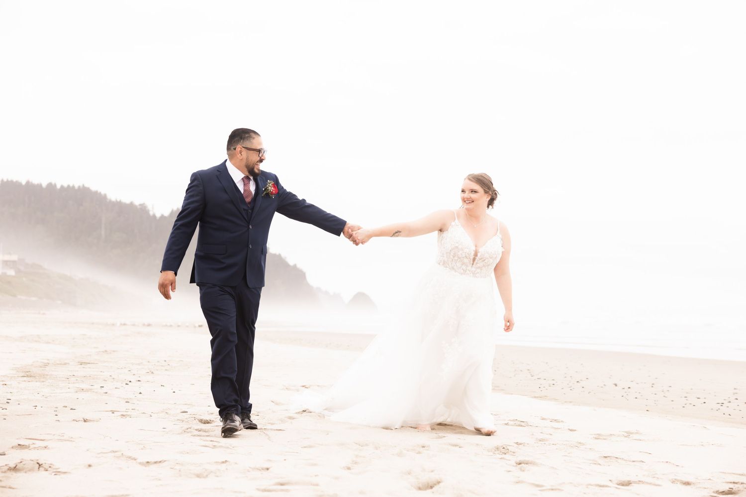 A couple in wedding attire holds hands while walking along a misty beach.