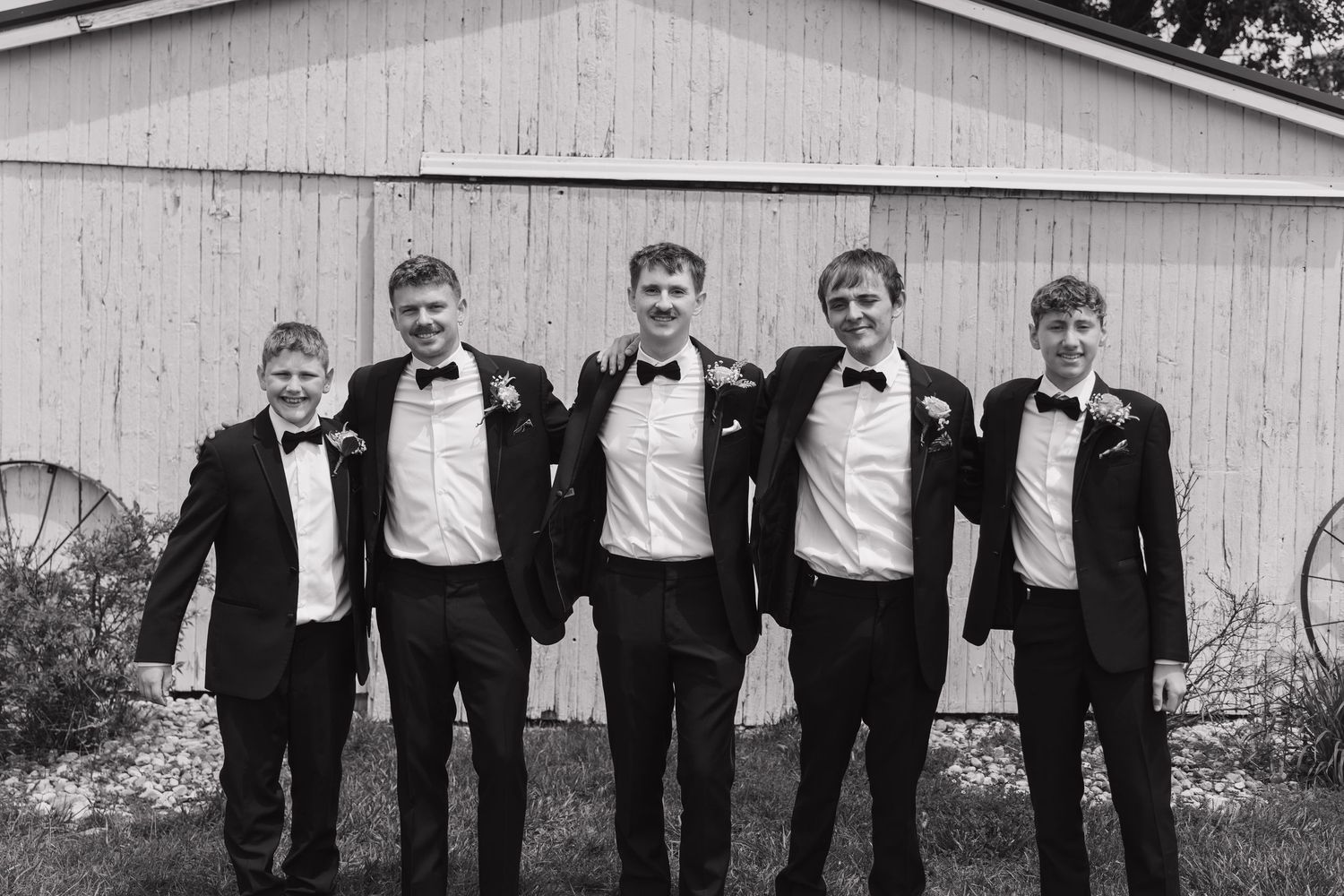 A group in formal black tuxedos and white shirts stand together in a line against a white building exterior.