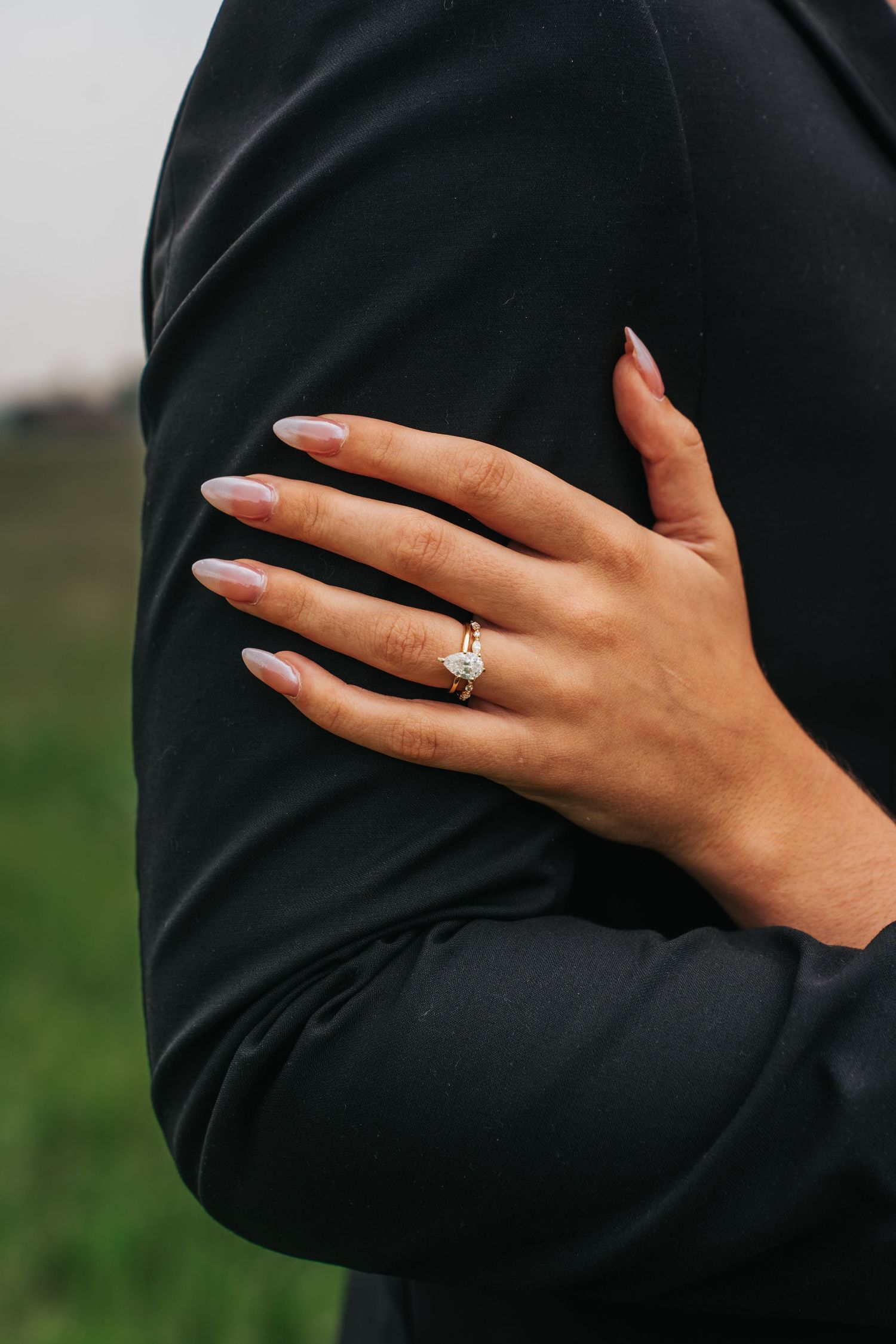 Close-up of a hand wearing an engagement ring resting against a black sleeve with green grass in background.