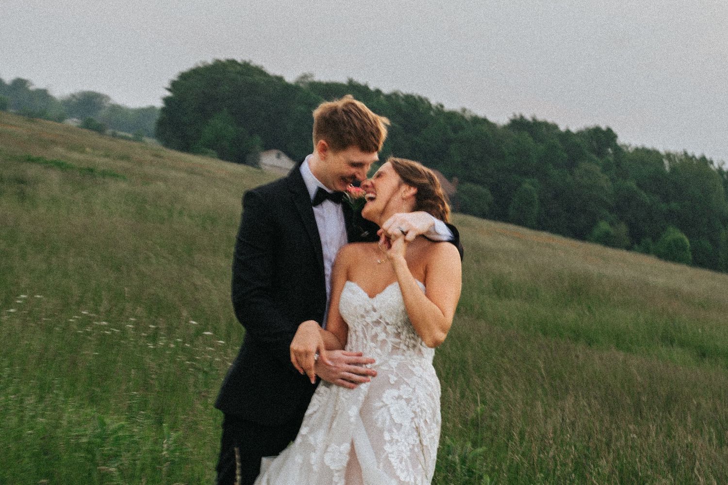 Couple in wedding attire embraces lovingly in a scenic meadow with rolling hills and trees in the background.