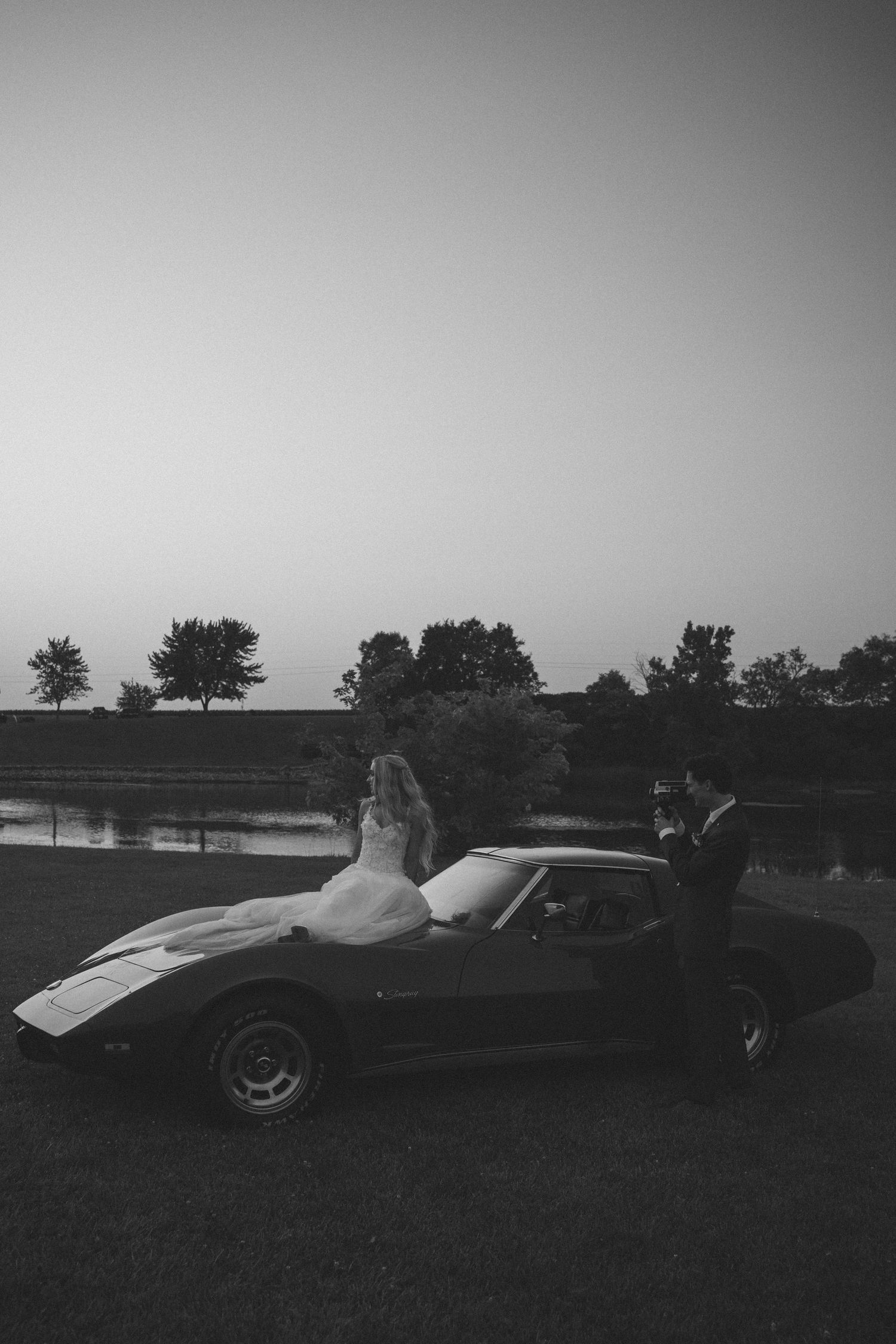 Black and white wedding photo of bride and groom posing beside a classic sports car at dusk near a serene lake.