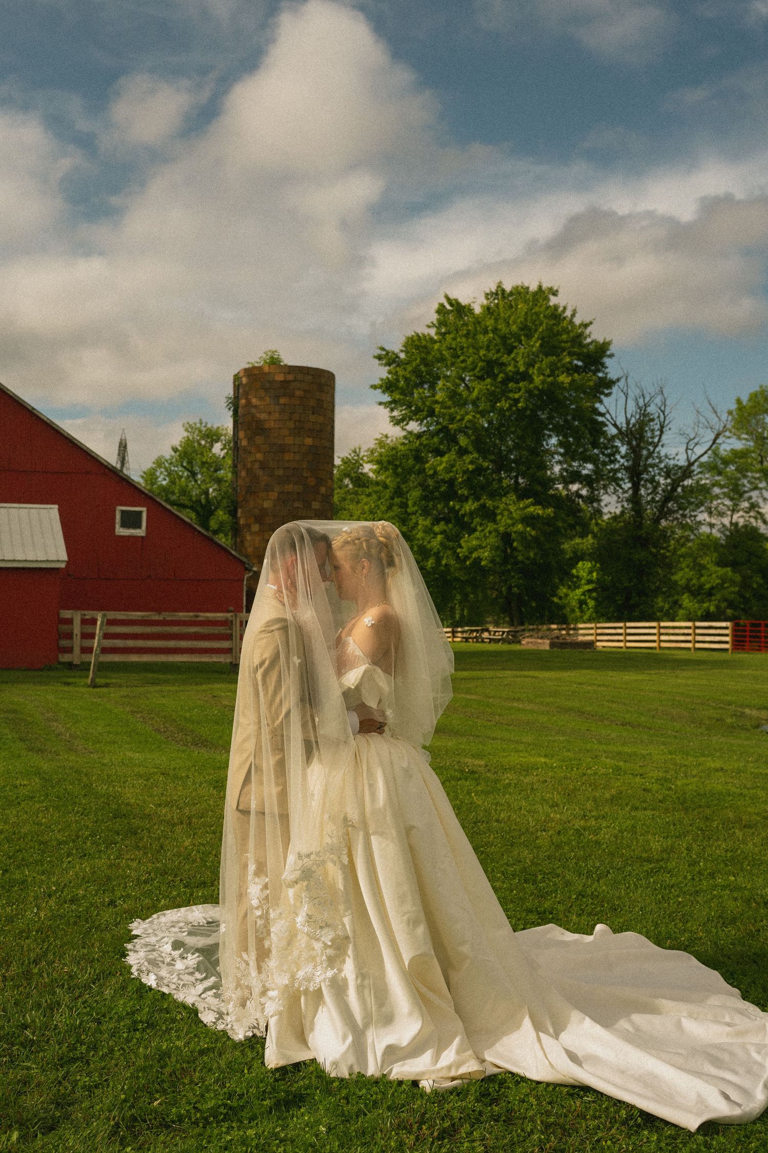 Bride in white gown and veil kissing groom outdoors near red barn and silo on lush green farm.