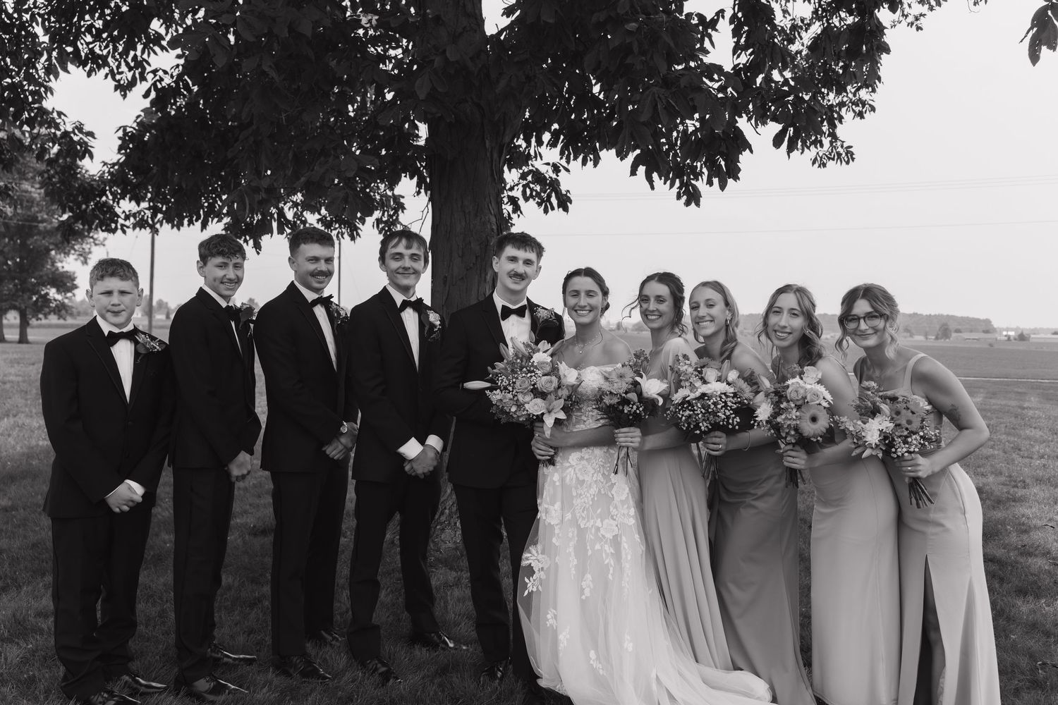 Wedding party members pose together outdoors in formal attire with bridesmaids in gray dresses and groomsmen in black suits.