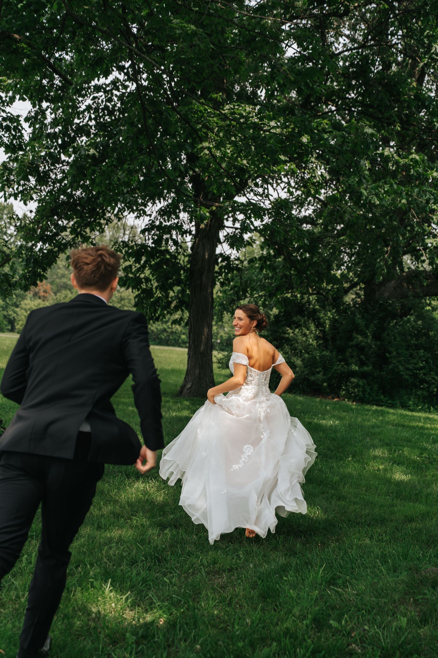 A couple in wedding attire playfully chase each other through a lush green garden setting.