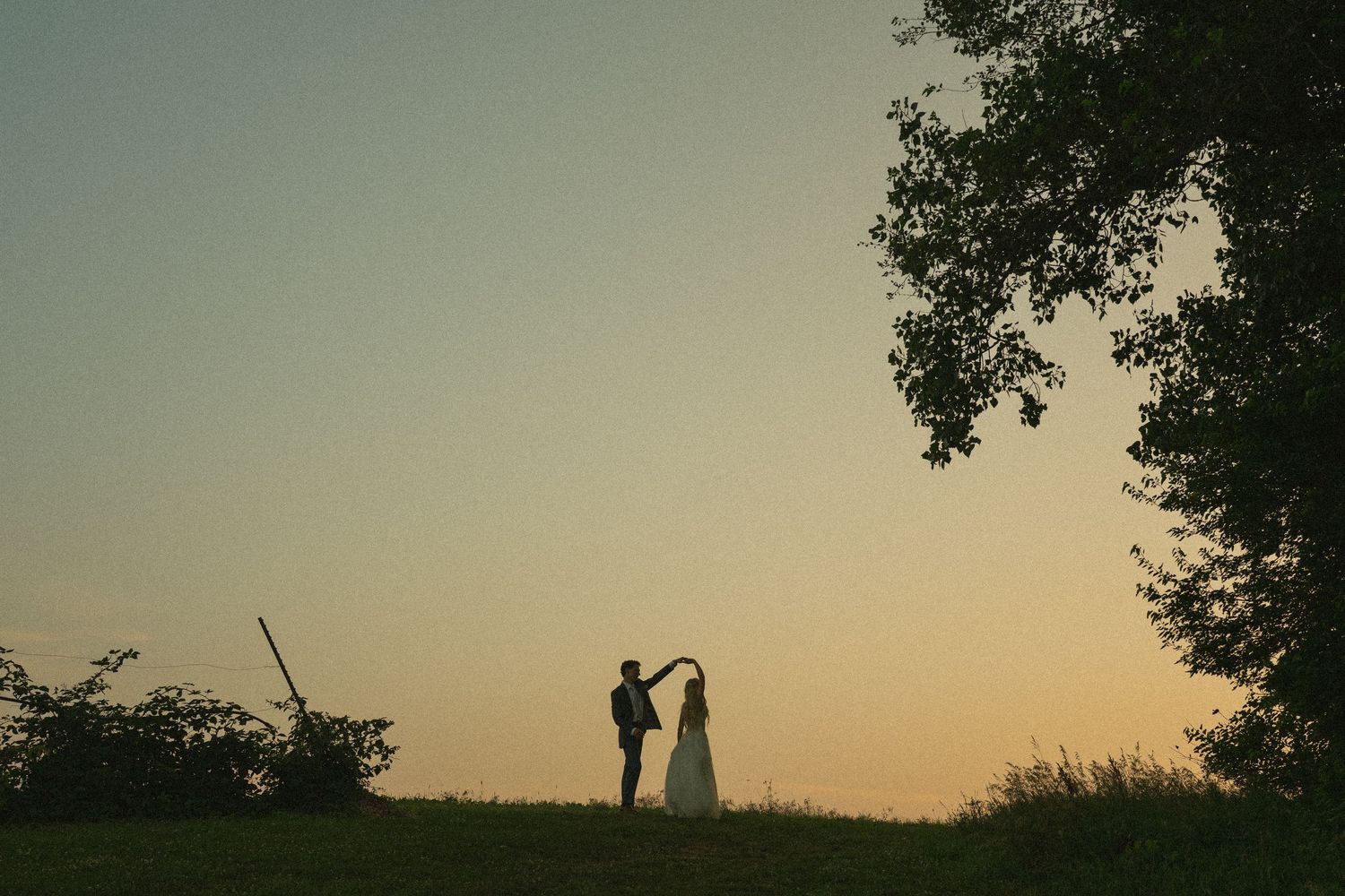 Silhouette of a couple dancing at sunset in a field, with a golden sky and trees framing the romantic scene.