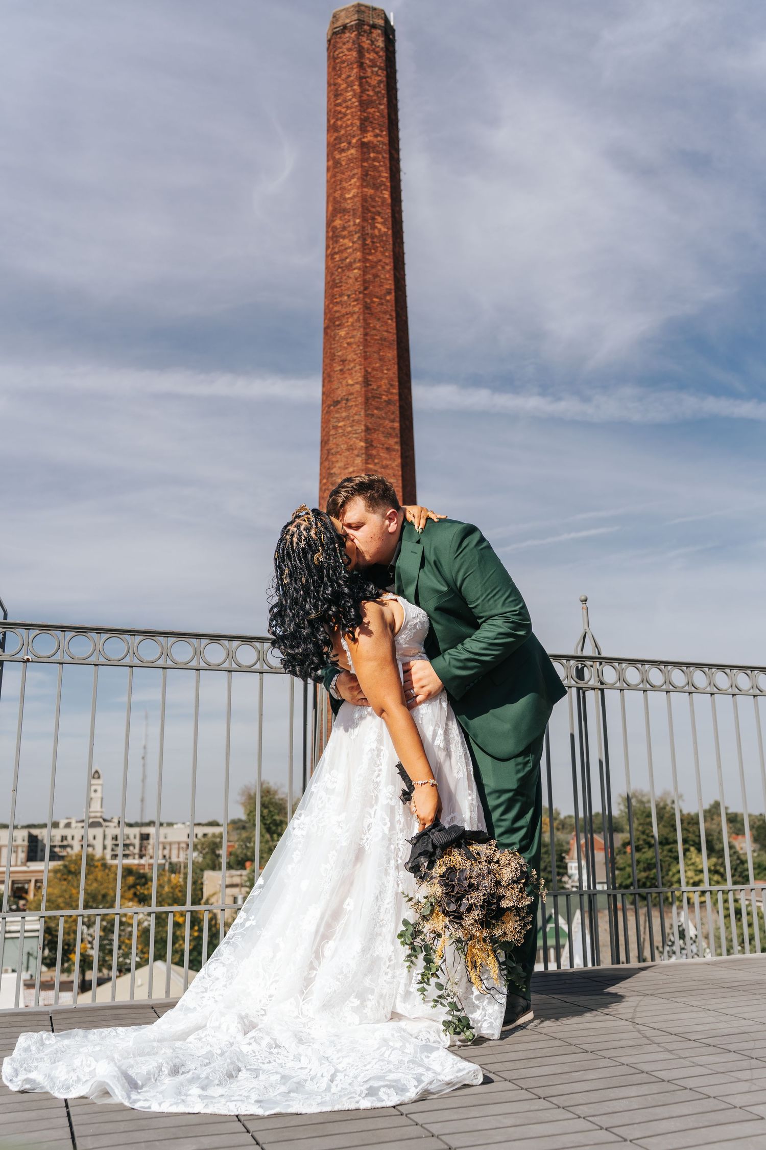 A couple in wedding attire share romantic kisses on a rooftop terrace with brick chimneys and metal railings.