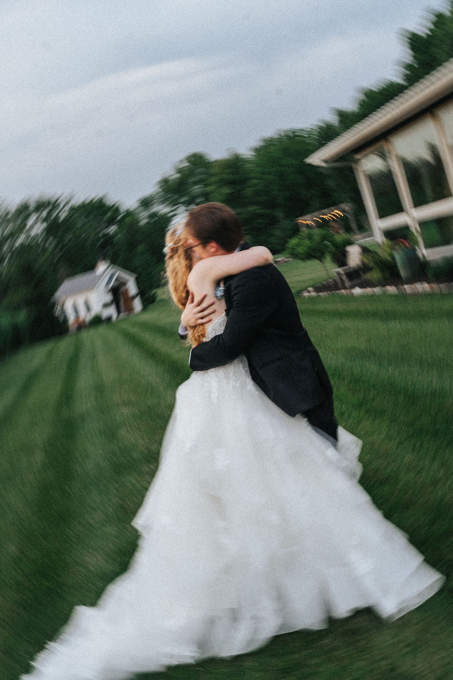 A couple in formal wedding attire spins and dances on a lush green lawn, creating a romantic motion-blur effect.