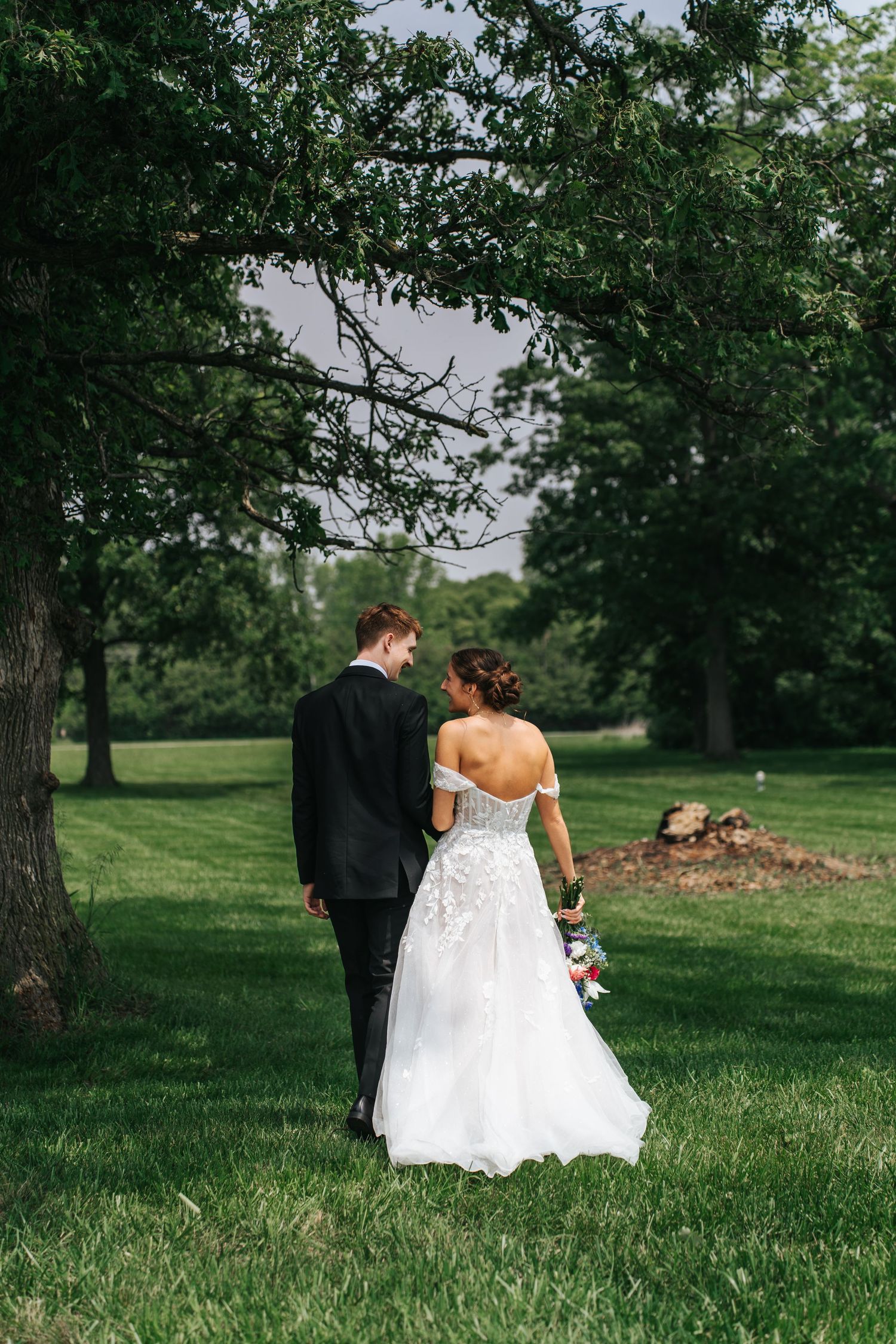 A bride and groom walk together across a lush green lawn surrounded by tall trees on their wedding day.