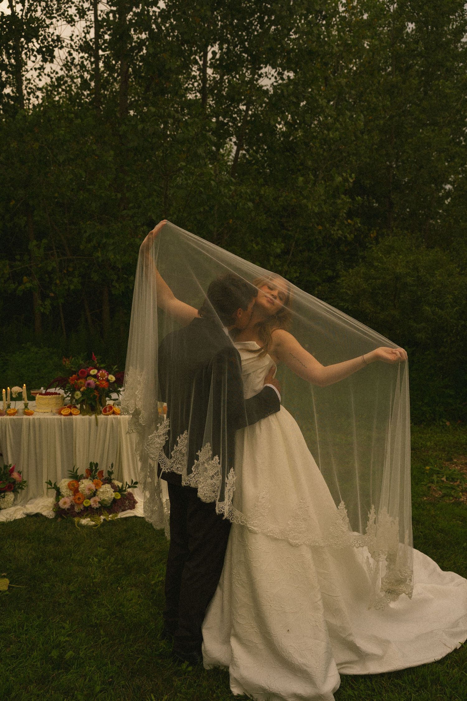 Bride and groom share romantic kiss under flowing lace veil at outdoor garden wedding reception.