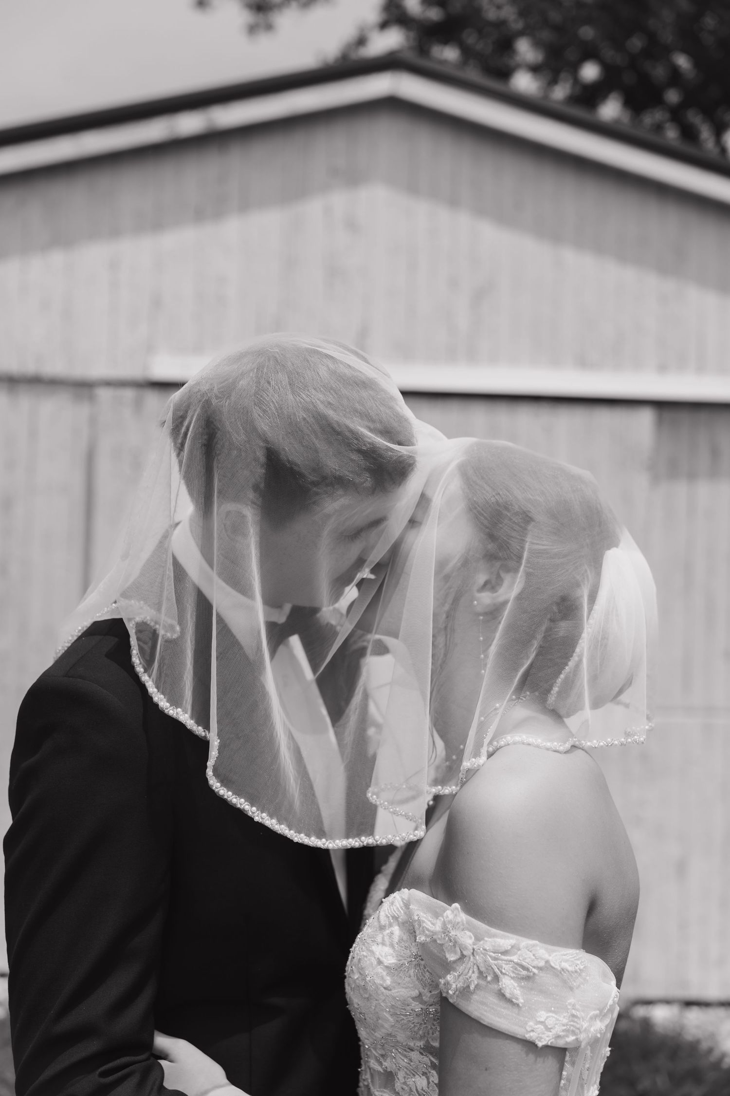 A romantic moment under a white veil as a couple shares a kiss in formal wedding attire.