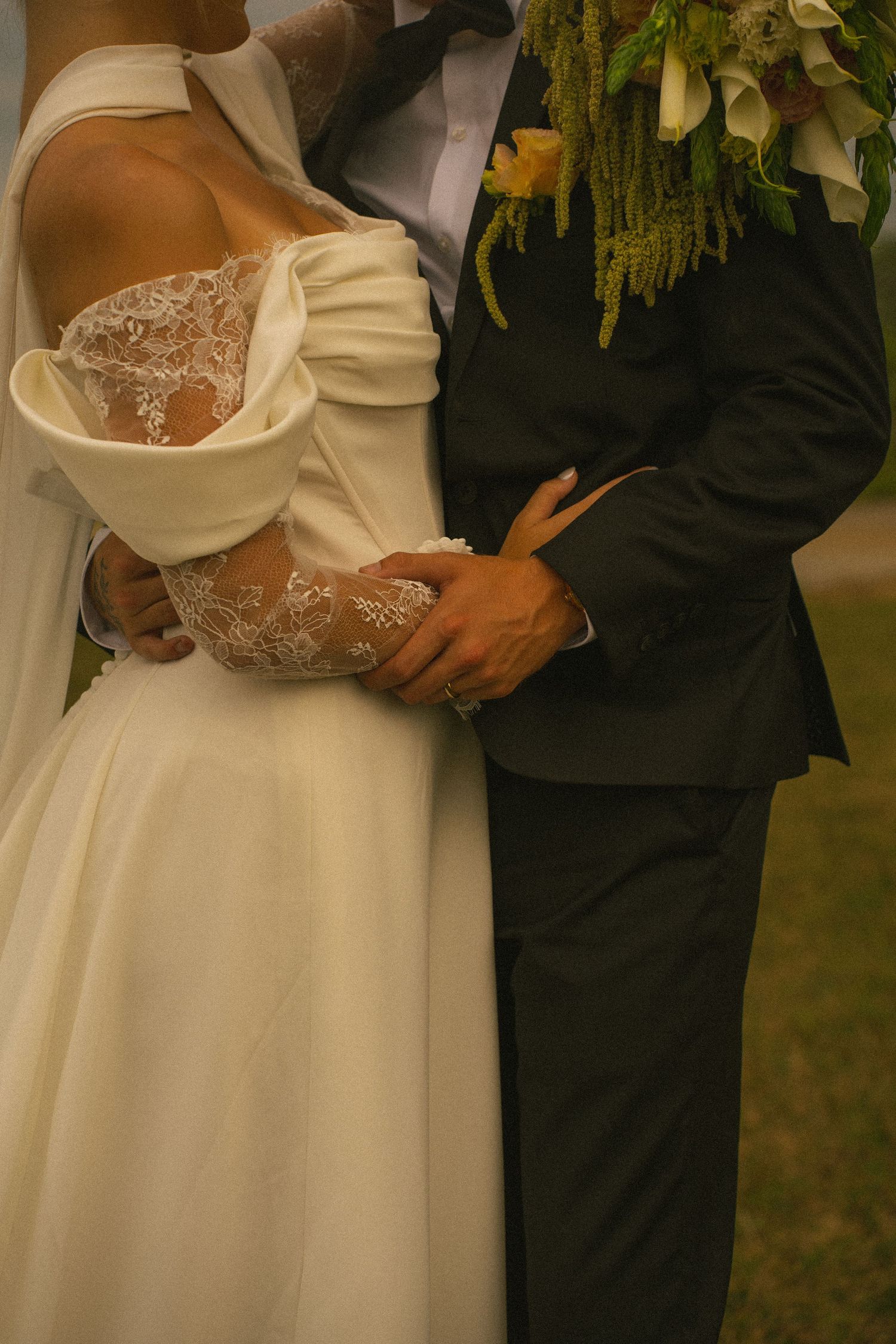 Bride in white off-shoulder lace gown embraced by groom in dark suit holding floral bouquet at wedding.